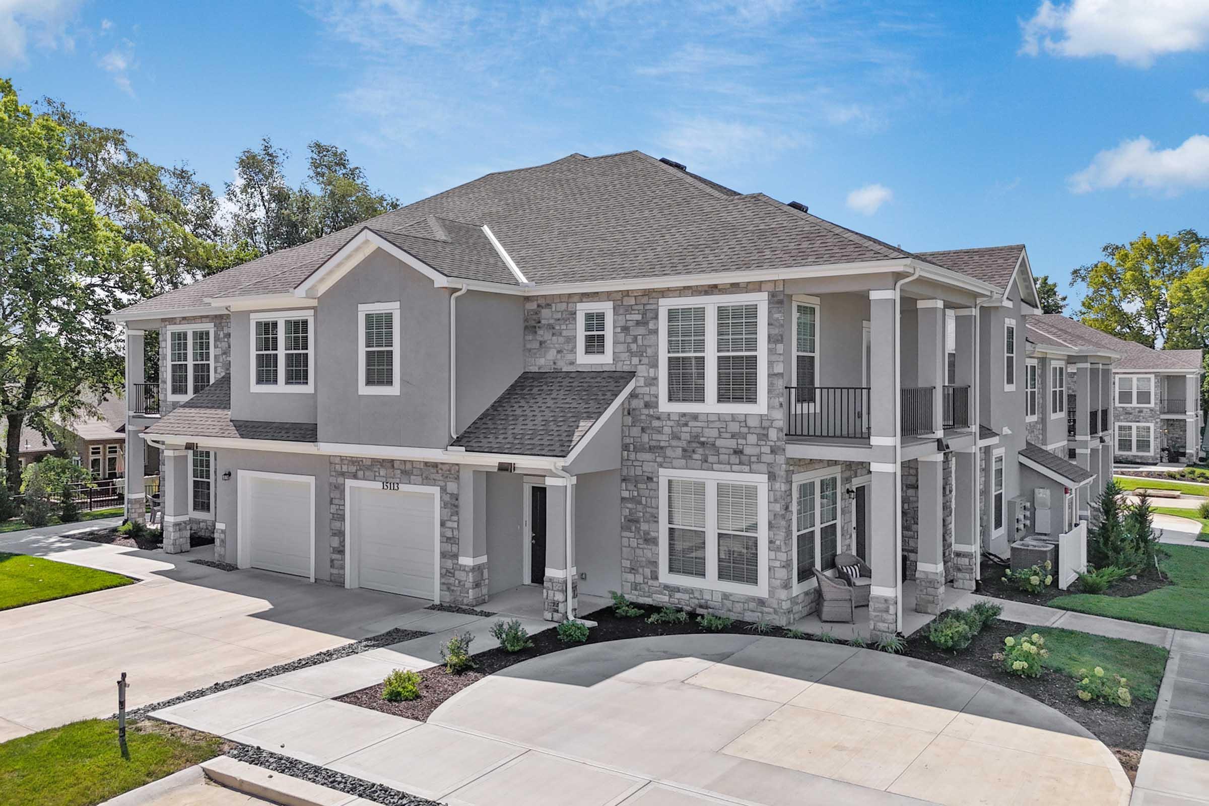 A modern two-story apartment building featuring a combination of gray siding and stone accents, with multiple balconies. The building has garages at the front and is surrounded by well-maintained landscaping, including green lawns and pathway decor. The sky is clear with a few clouds.