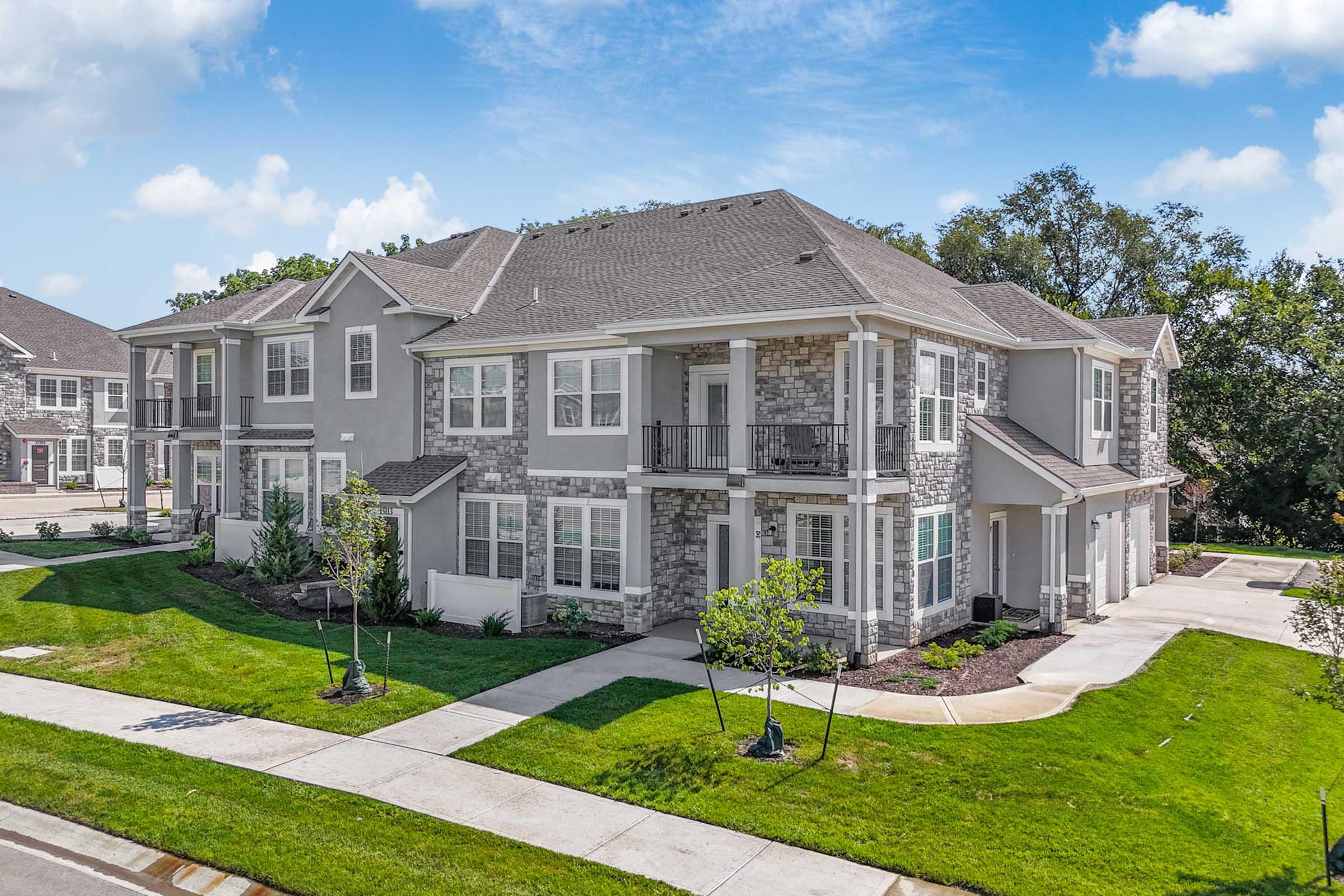 A view of a modern residential building featuring a mix of stone and siding exteriors with multiple balconies. Lush green landscaping surrounds the property, and a well-maintained sidewalk leads to the entrance. The setting is sunny with blue skies in the background.