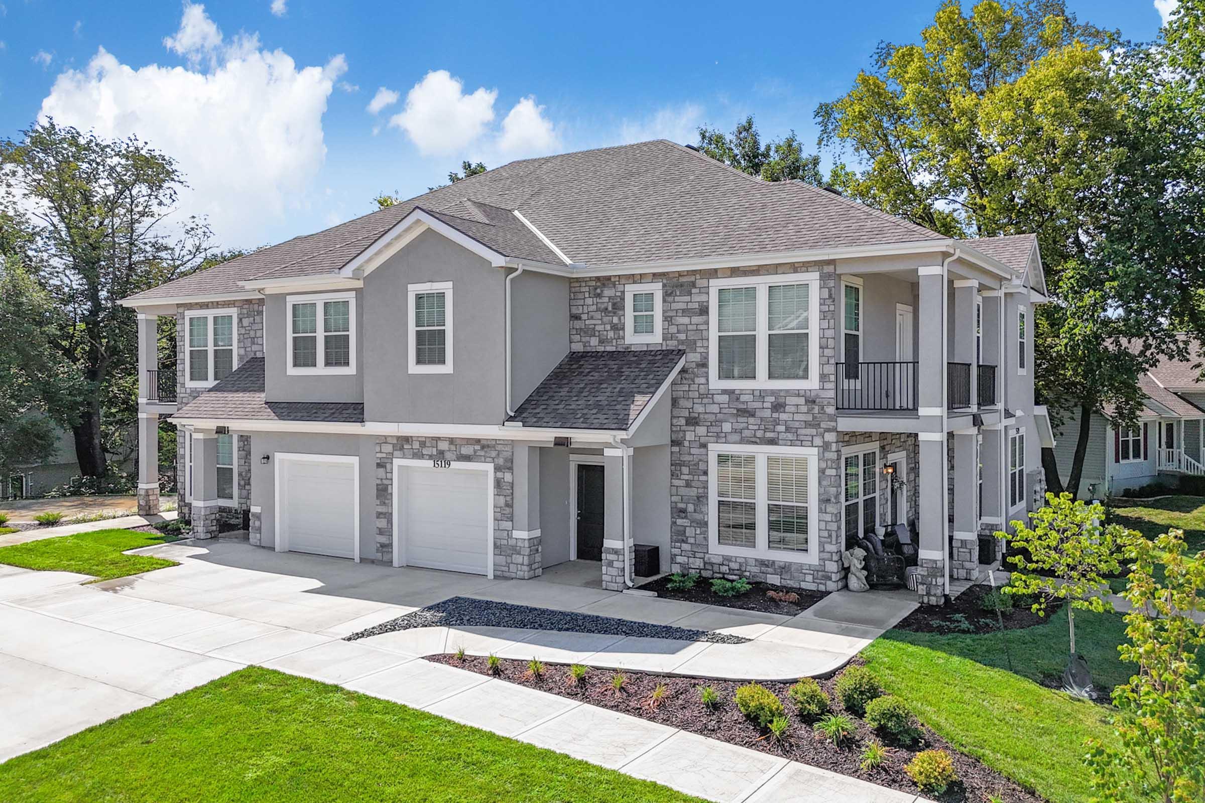 A large, modern two-story house featuring a combination of gray siding and stone accents. It has a spacious front yard with landscaped gardens, a driveway leading to a two-car garage, and several windows that offer natural light. Trees and blue sky in the background complete the picturesque scene.