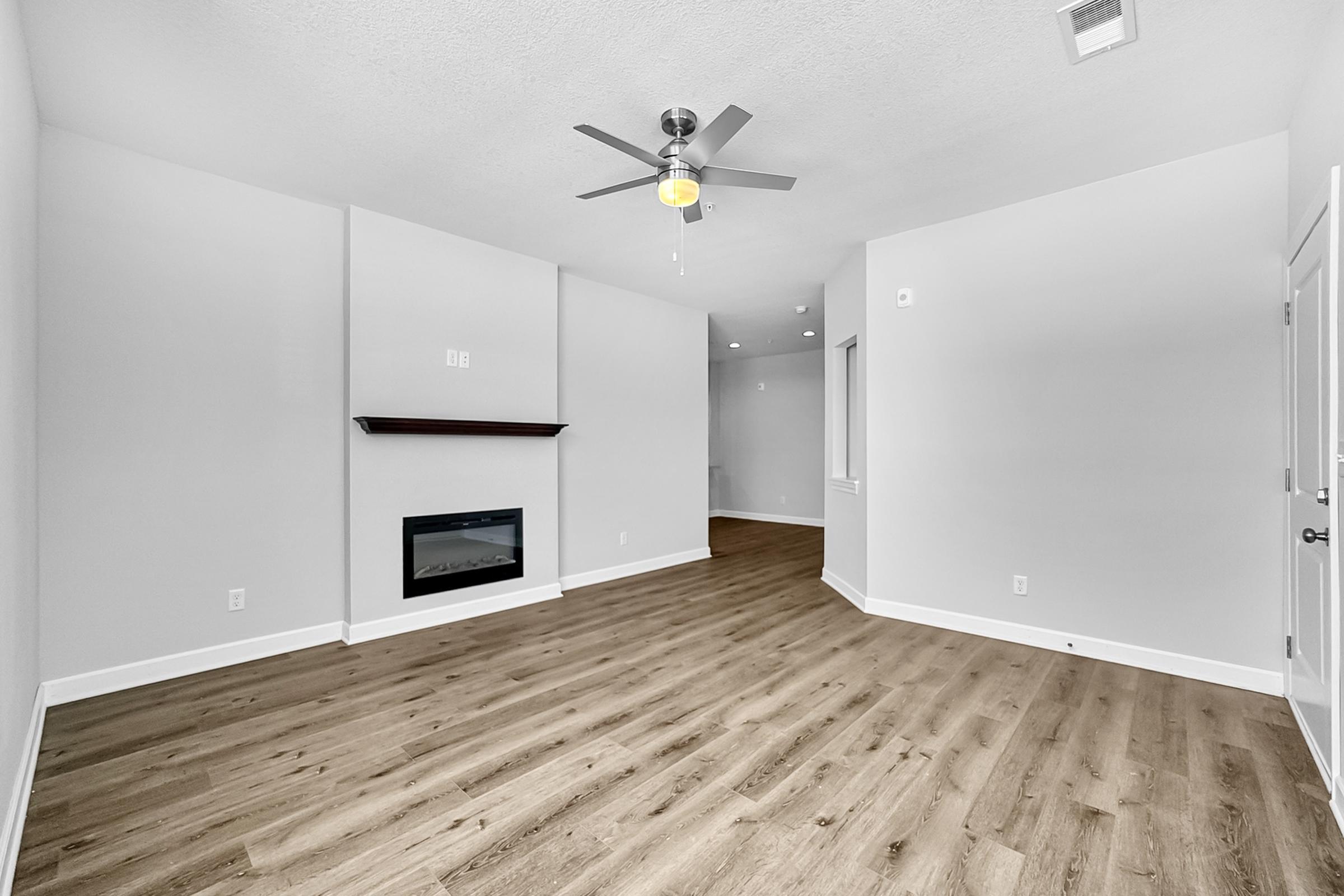 A spacious, empty living room with light gray walls and wood-like flooring. It features a modern ceiling fan and a simple electric fireplace in the center. Natural light comes through a window on the right, and there are no furnishings, creating an open and inviting atmosphere.