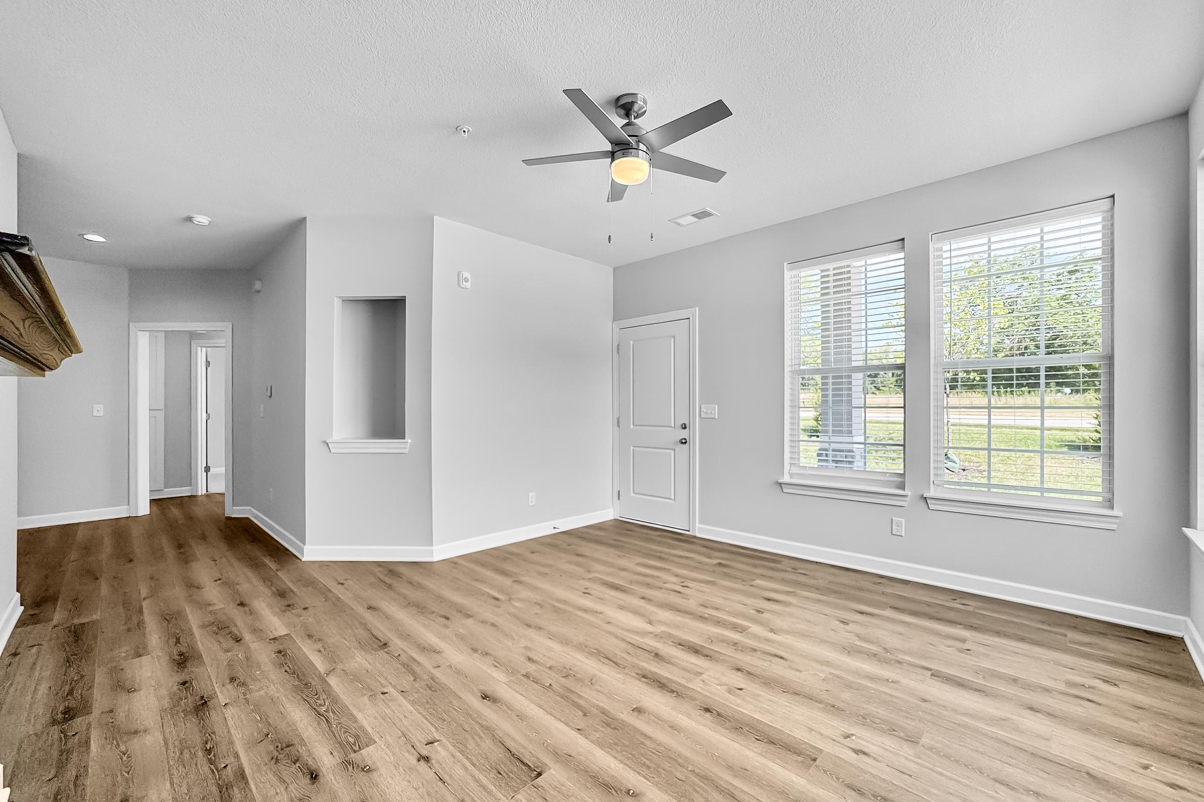 Spacious, empty living room with light gray walls, large windows allowing natural light, and wooden flooring. A ceiling fan is mounted in the center. A doorway leads to another room, and there is a small alcove near the entrance. The atmosphere is bright and airy.