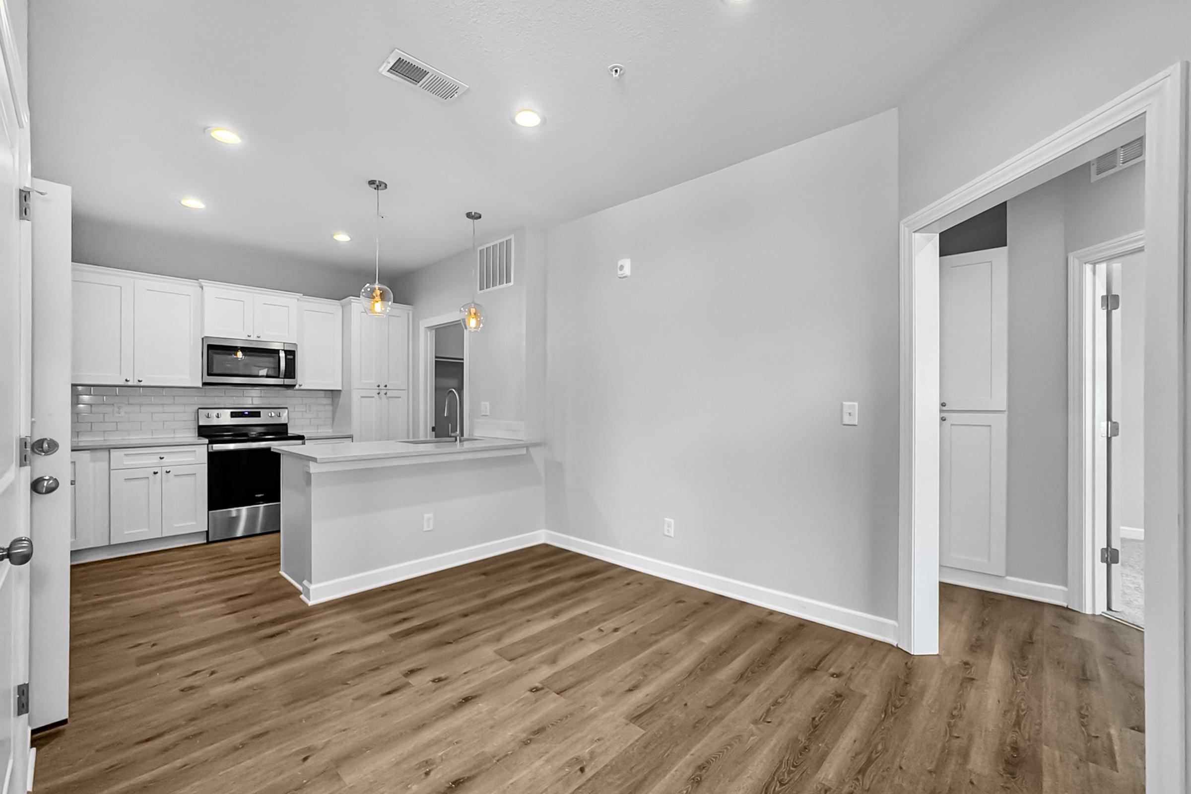 A modern kitchen and living area featuring white cabinets, stainless steel appliances, and an open layout. The space has warm wood-like flooring and recessed lighting, with a counter bar area and a doorway leading to a hallway. The walls are painted a light gray, creating a bright and airy atmosphere.