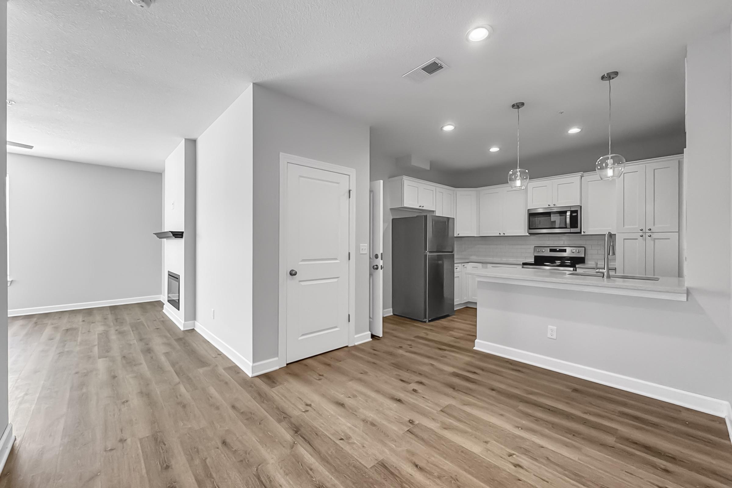 Modern open-concept living space featuring a kitchen with stainless steel appliances, a white cabinetry, and a small dining area. Light-colored walls and wood flooring create a bright and inviting atmosphere. A fireplace is visible in the living area, adding a cozy touch.