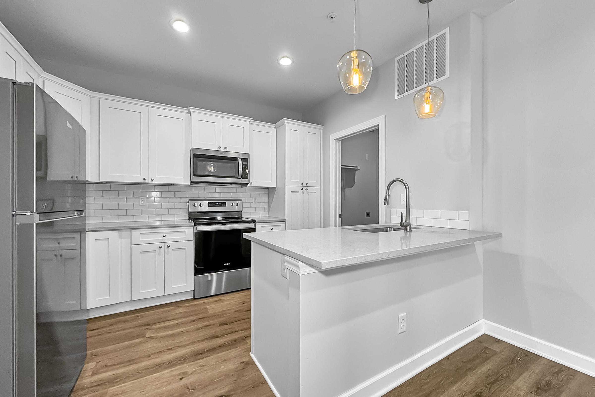 Modern kitchen featuring white cabinetry, stainless steel appliances, and quartz countertops. The space includes a kitchen island with a sink and pendant lighting, along with a light-colored backsplash. Wood-like flooring adds warmth to the contemporary design.