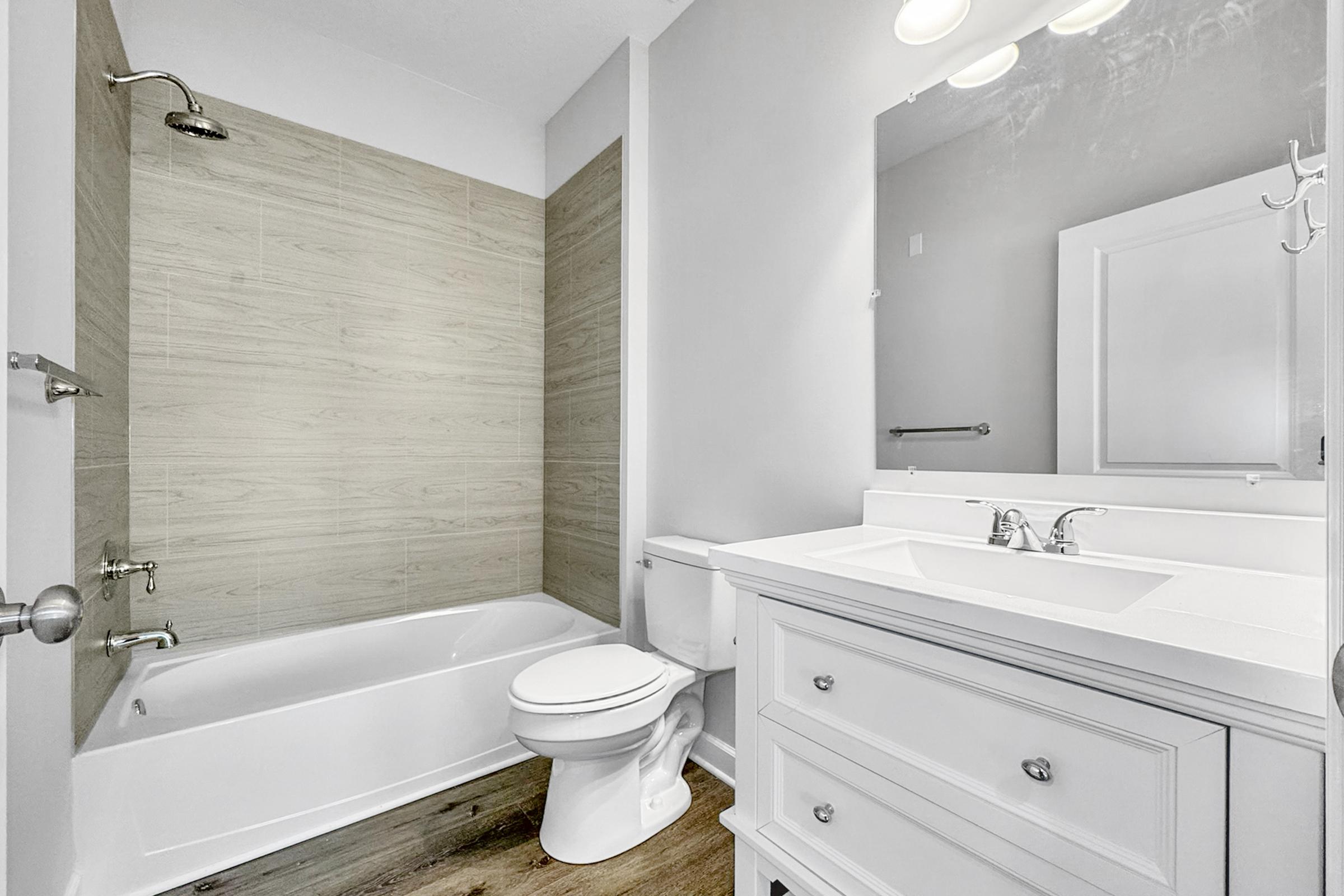A modern bathroom featuring a bathtub with a shower, light-colored wall tiles, and a white vanity with a sink. There is a large mirror above the vanity, a toilet, and neutral-colored flooring. The overall design is bright and contemporary.
