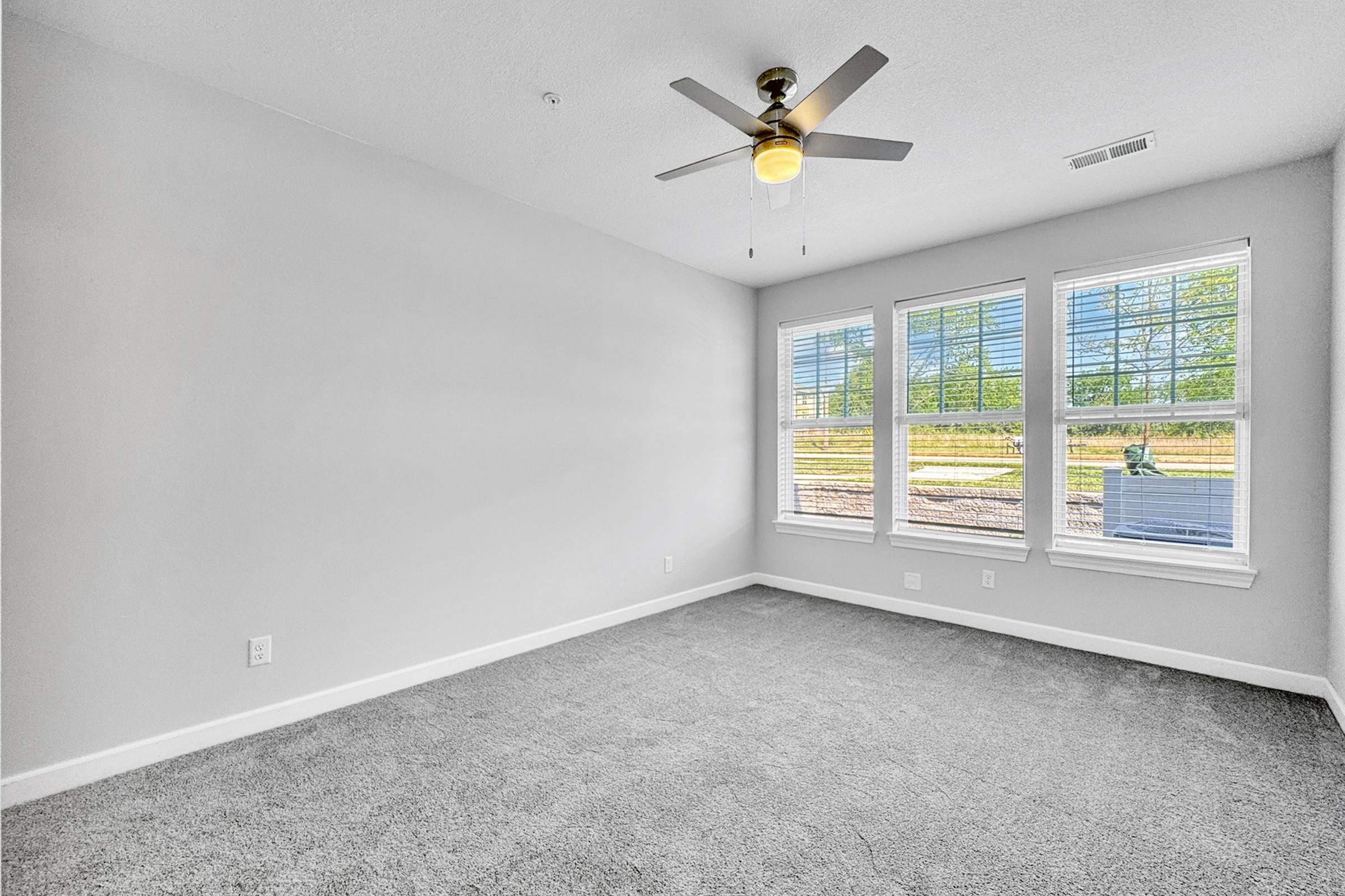 A bright, empty room with light gray carpet, featuring a ceiling fan with a light fixture. There are three large windows on one wall, allowing natural light to fill the space. The walls are painted a light color, and the room has a clean, modern aesthetic.