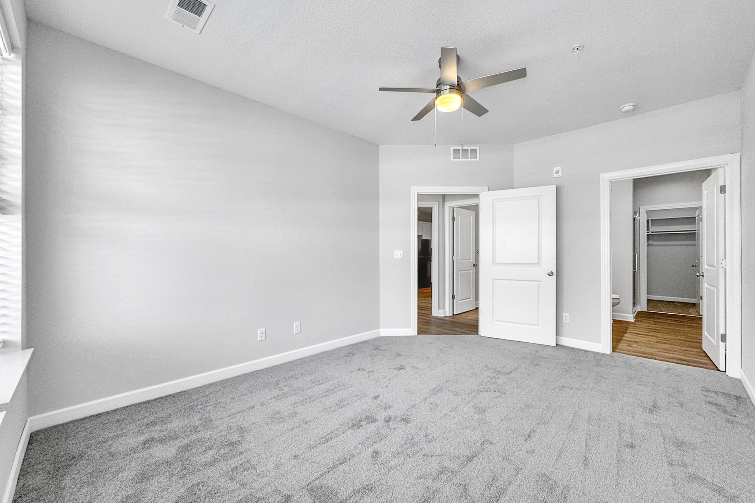 A spacious, light-filled room with light gray walls and plush carpet. It features a ceiling fan and has a large window allowing natural light. Doors lead to a closet and adjacent rooms. The overall atmosphere is modern and inviting, ideal for a bedroom or study.
