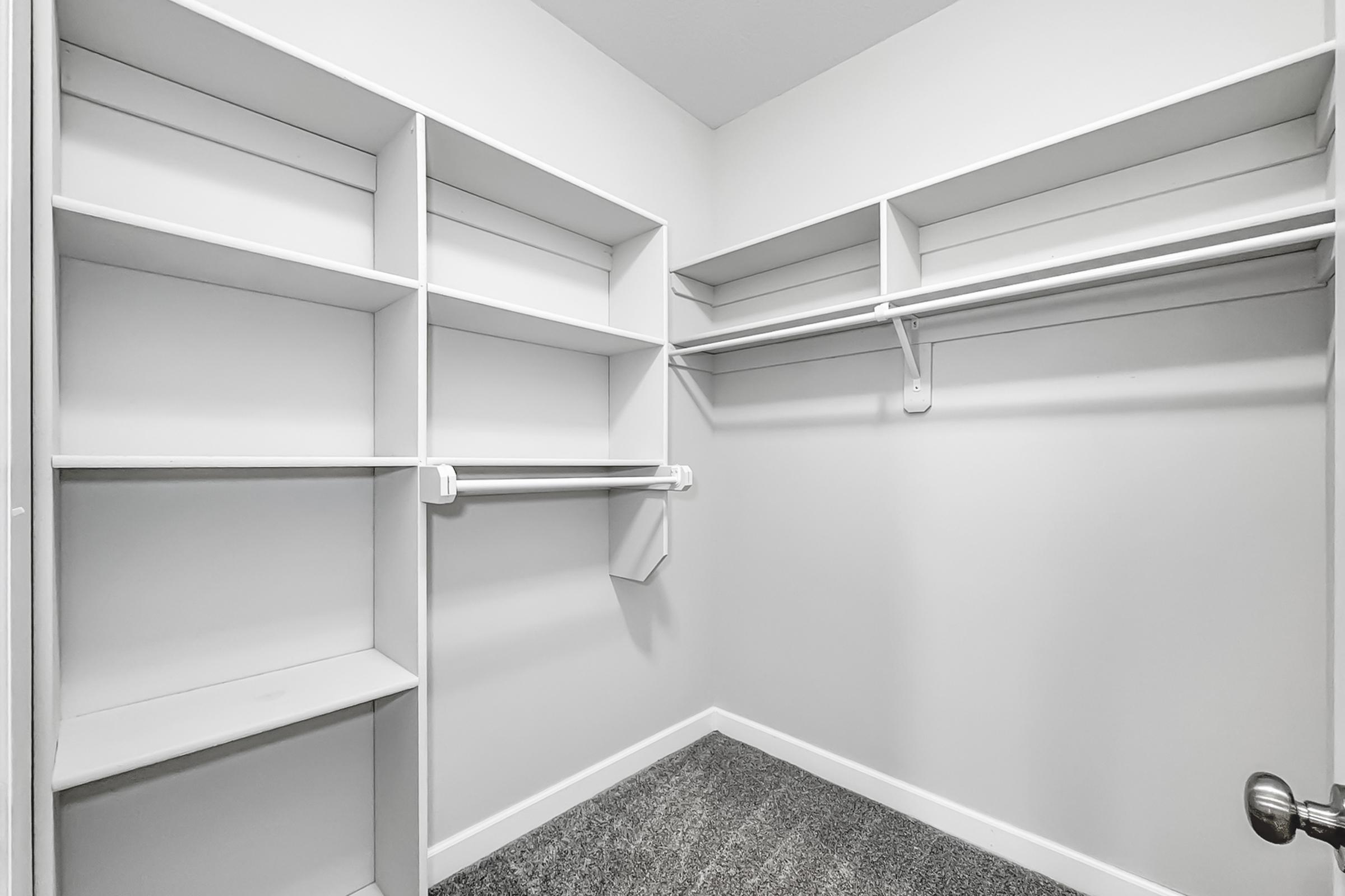 A spacious empty closet featuring white shelving units and hanging rods, with light gray carpet flooring. The walls and shelves are clean and minimalist, creating an organized look. Ideal for storage or organization.