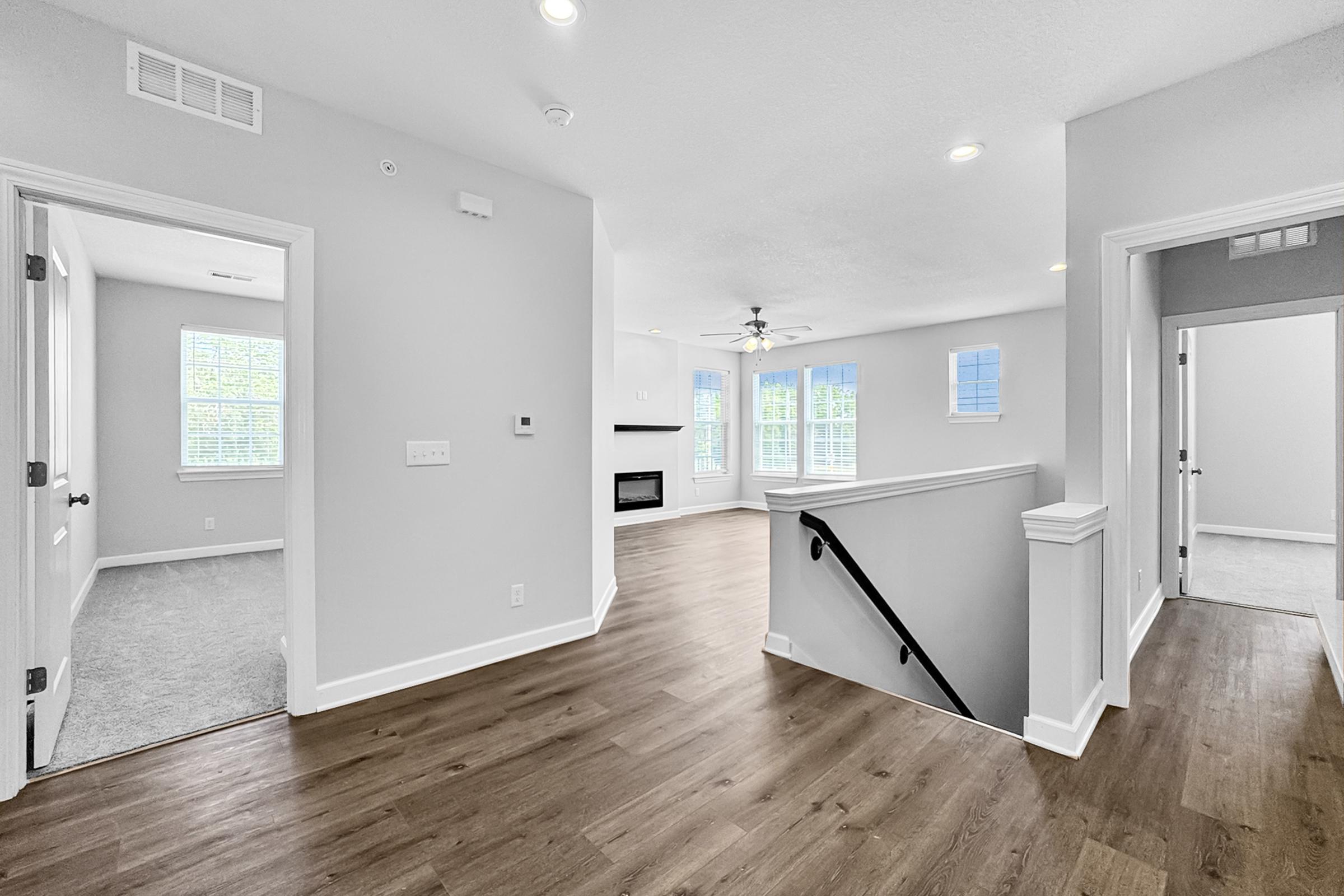 Bright, spacious interior of a modern home featuring light gray walls, a ceiling fan, and large windows. The open layout includes a living area with a fireplace, and a staircase leading to an upper level. The floors are a warm, wood-like finish, with plush carpet in the adjacent room.