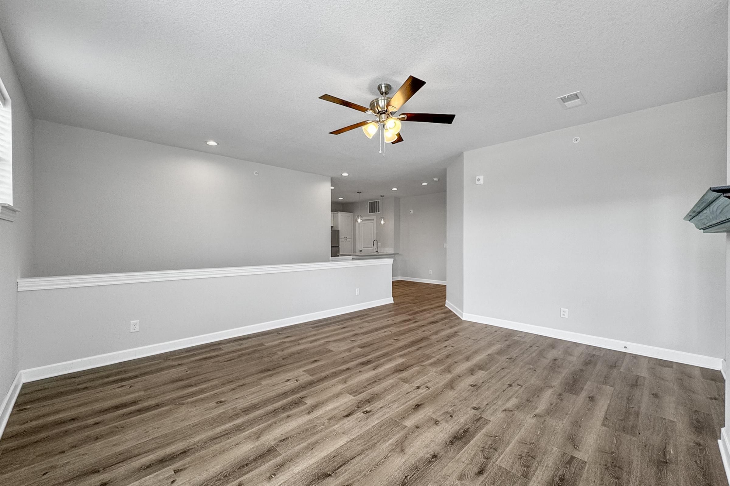 A spacious, well-lit living area featuring light gray walls and a ceiling fan. The floor is adorned with wood-like laminate, and there are large windows allowing natural light. In the background, an open kitchen area can be seen, creating an inviting and modern atmosphere.