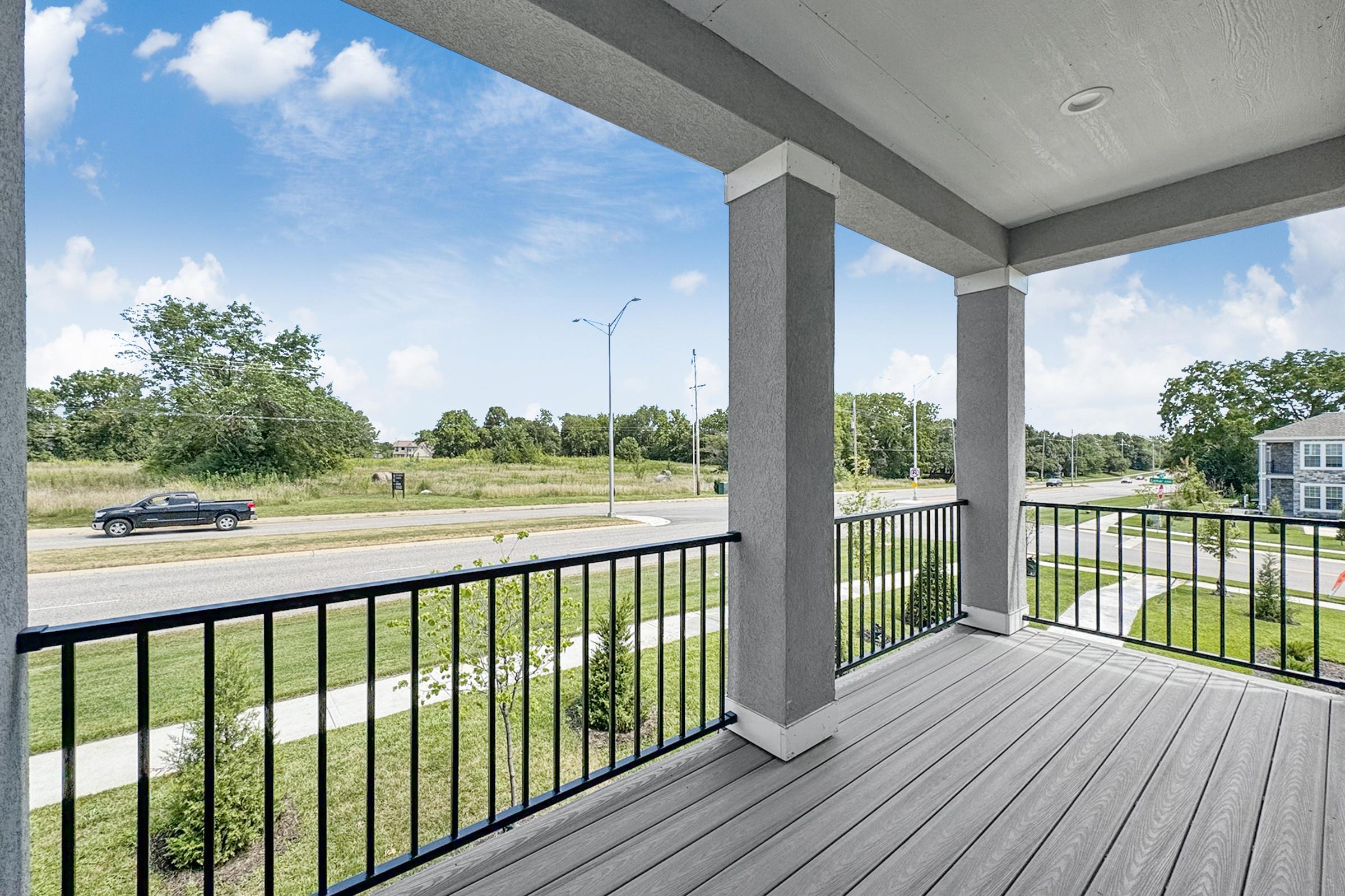 A view from a balcony overlooking a landscaped area and a road. The scene includes green grass and trees, with a car driving by in the distance. The sky is bright with fluffy clouds. The balcony features a railing and composite flooring, creating a spacious outdoor space.