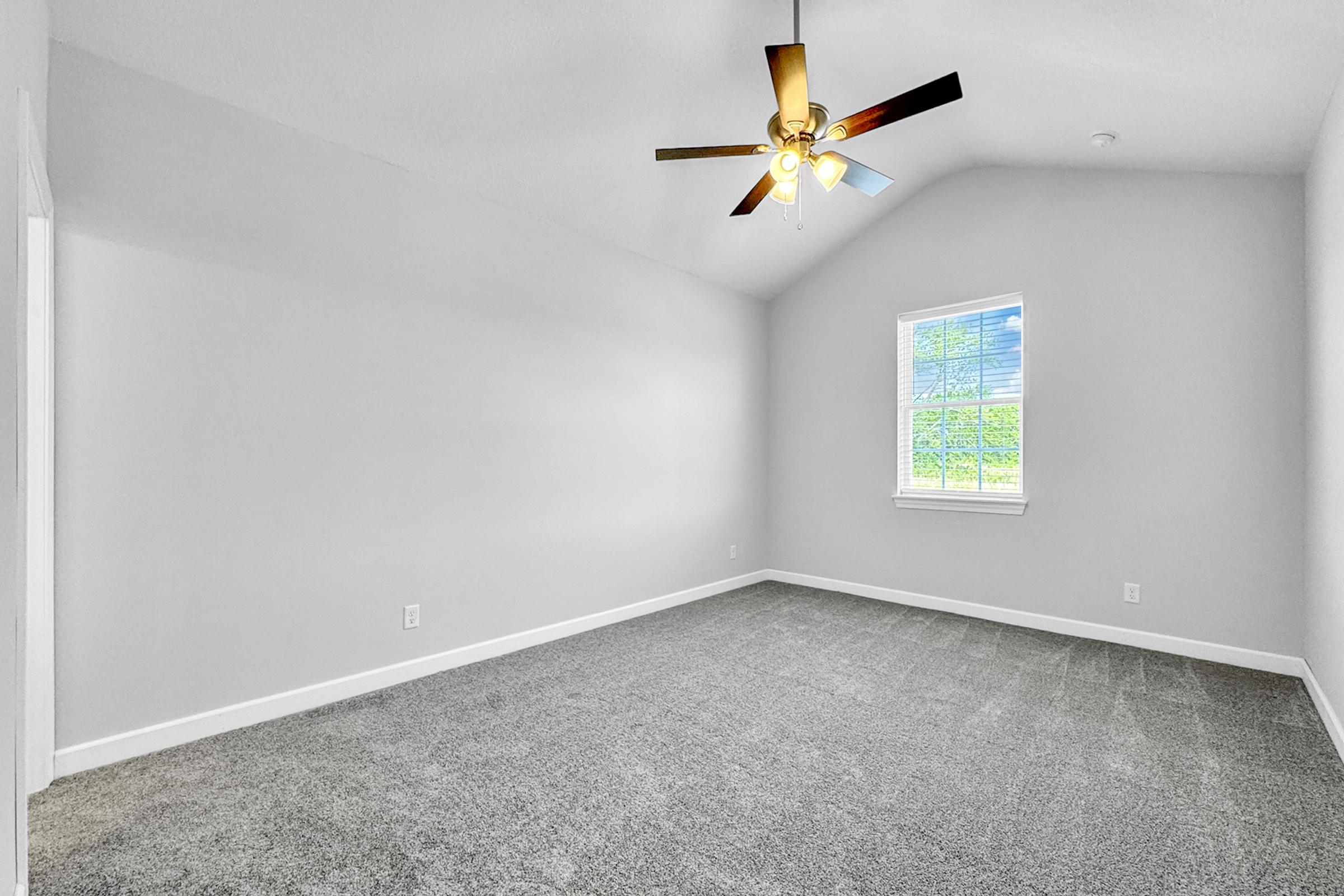 A spacious, empty room with light gray walls and plush carpet. The ceiling is high, featuring a ceiling fan with a light. A single window allows natural light to illuminate the space, which has a bright and airy feel.