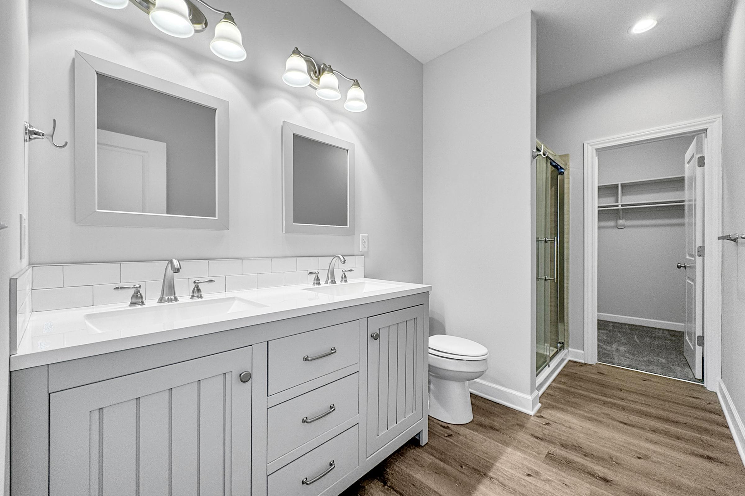 Modern bathroom featuring a dual sink vanity with gray cabinetry, two mirrors above the sinks, and polished chrome faucets. The space has light-colored walls, wooden flooring, and a glass shower enclosure. A closet is visible in the corner, adding to the functionality of the room.