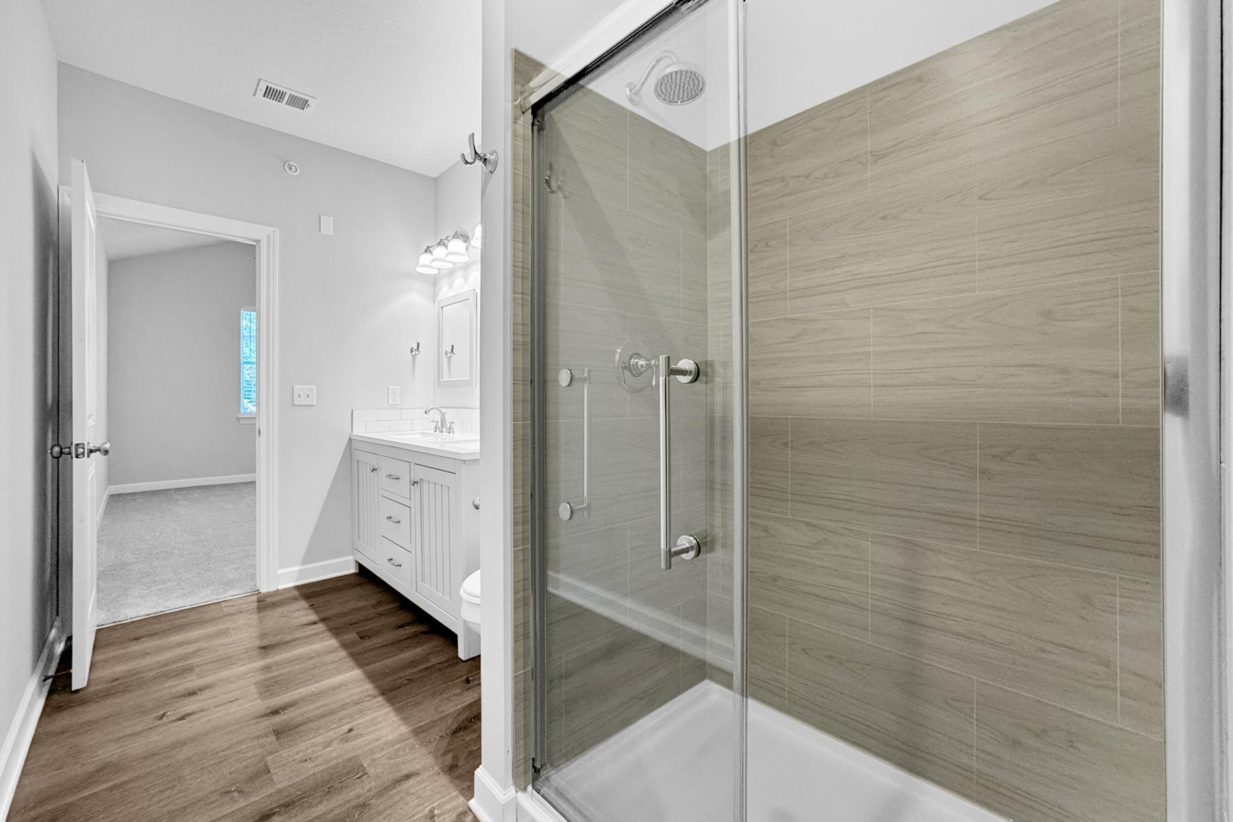 A modern bathroom featuring a glass shower enclosure with a neutral tile design, a white vanity with a sink, and light wood flooring. A doorway leads to another room, with soft natural light coming through the window. The overall design is clean and contemporary.