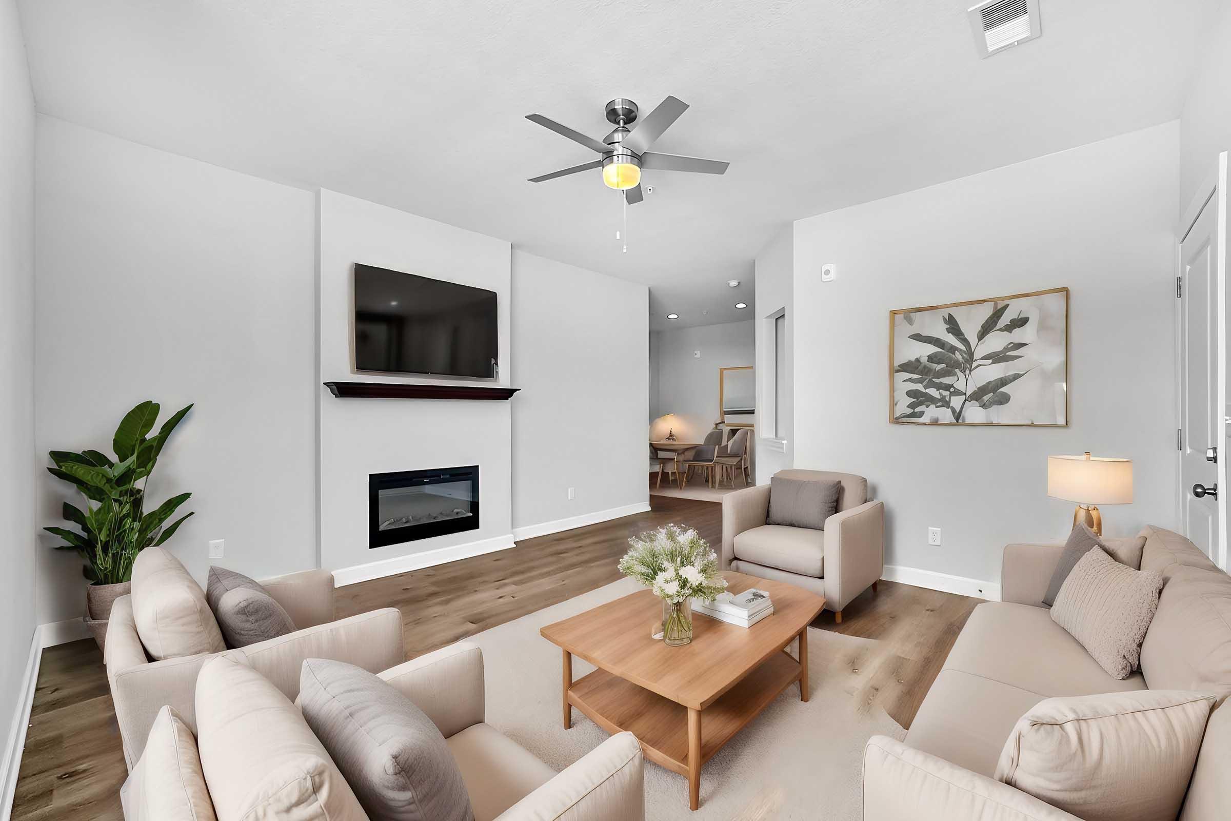 A modern living room featuring light-colored walls, hardwood flooring, and comfortable beige sofas arranged around a wooden coffee table. A flat-screen TV is mounted above a simple electric fireplace, and a large potted plant adds a touch of greenery. Soft lighting and a framed botanical print enhance the inviting atmosphere.