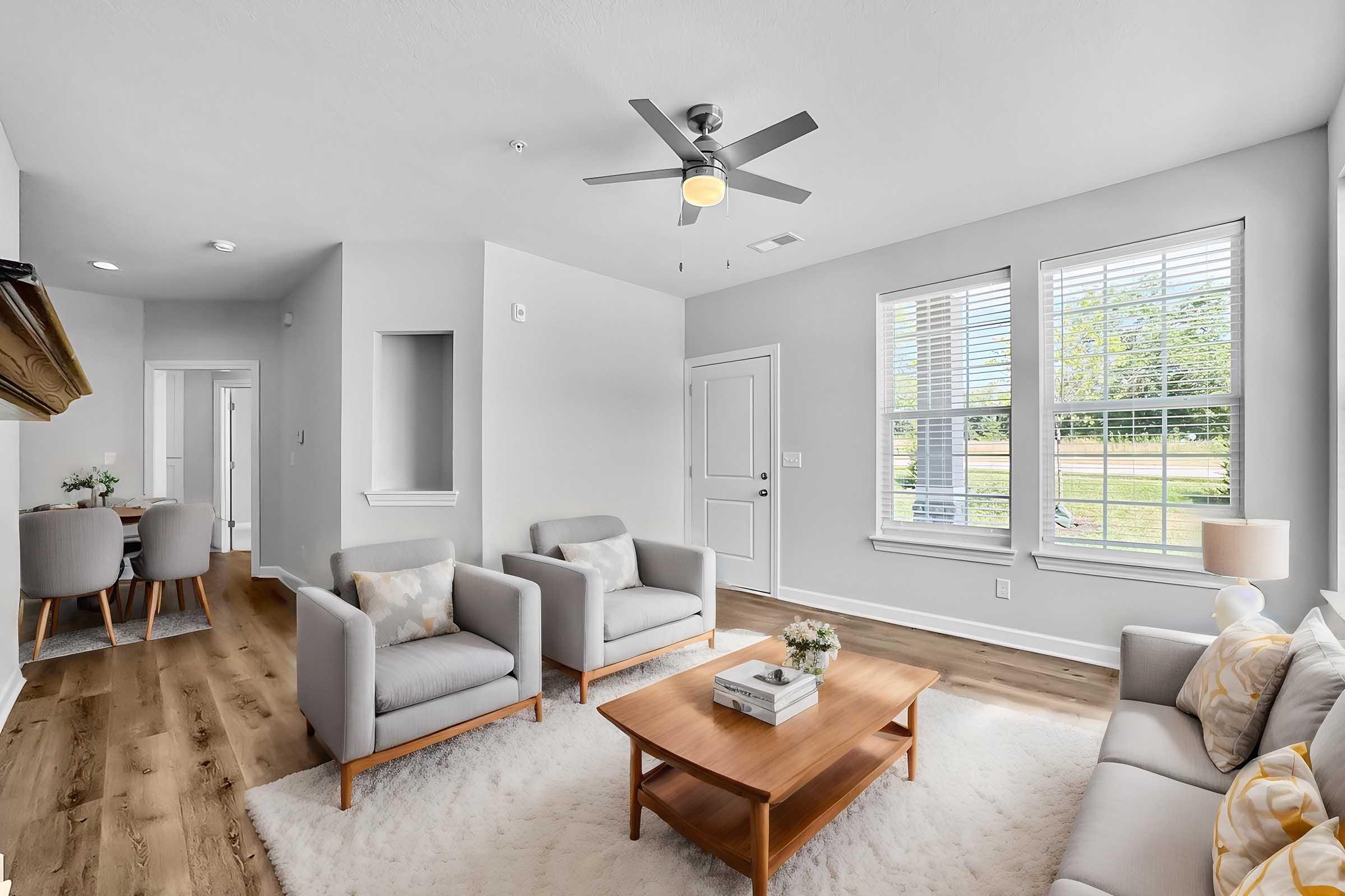 A bright and modern living room featuring light-colored walls, a ceiling fan, and hardwood floors. The room includes two armchairs and a sofa with decorative pillows around a wooden coffee table. Natural light streams in through large windows, enhancing the open and airy feel of the space, which connects to a dining area in the background.