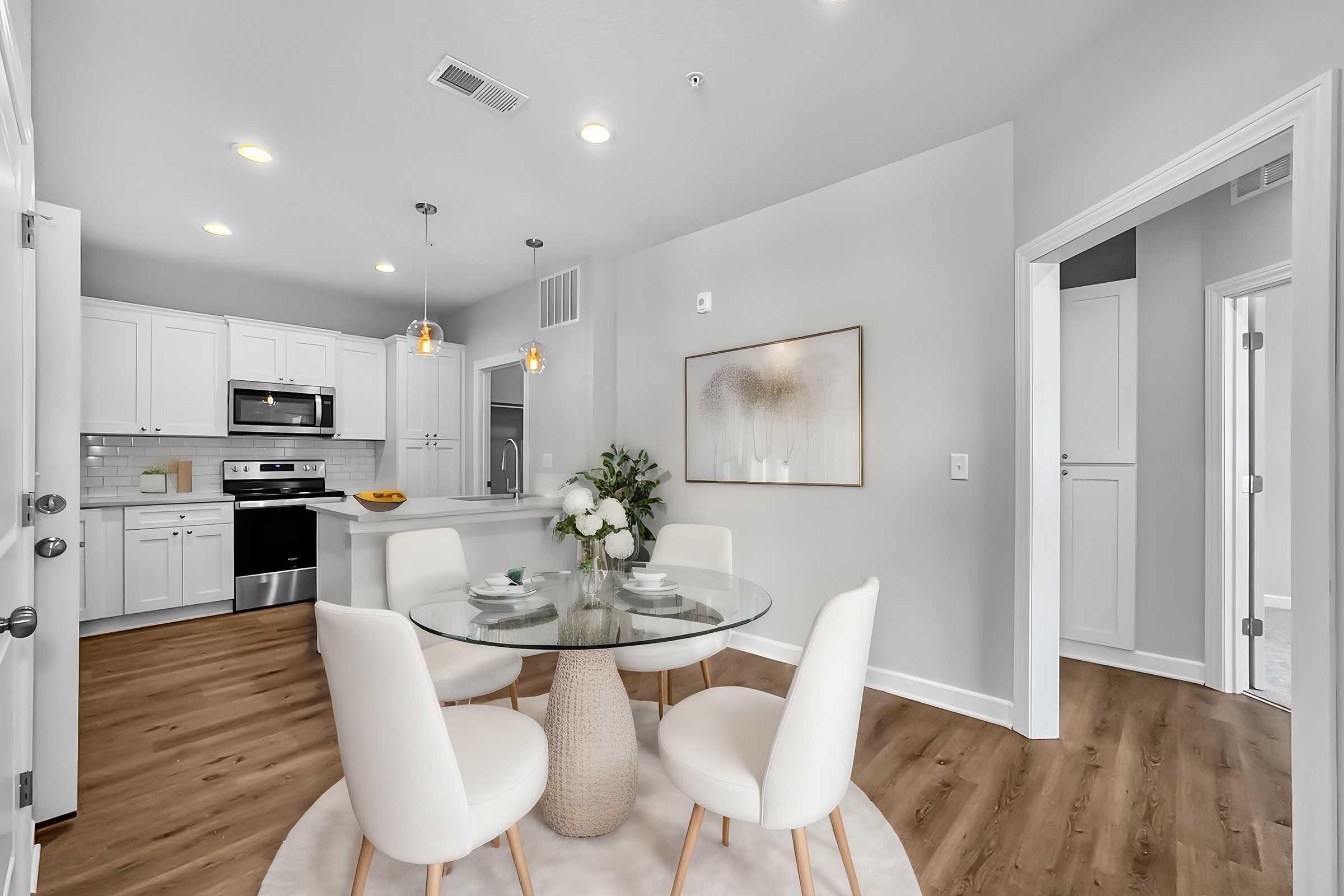 A modern kitchen and dining area featuring a round glass table with four white chairs, a floral centerpiece, and light wood flooring. The kitchen showcases white cabinets, stainless steel appliances, and warm lighting. A doorway leads to another room, creating an inviting and stylish space.