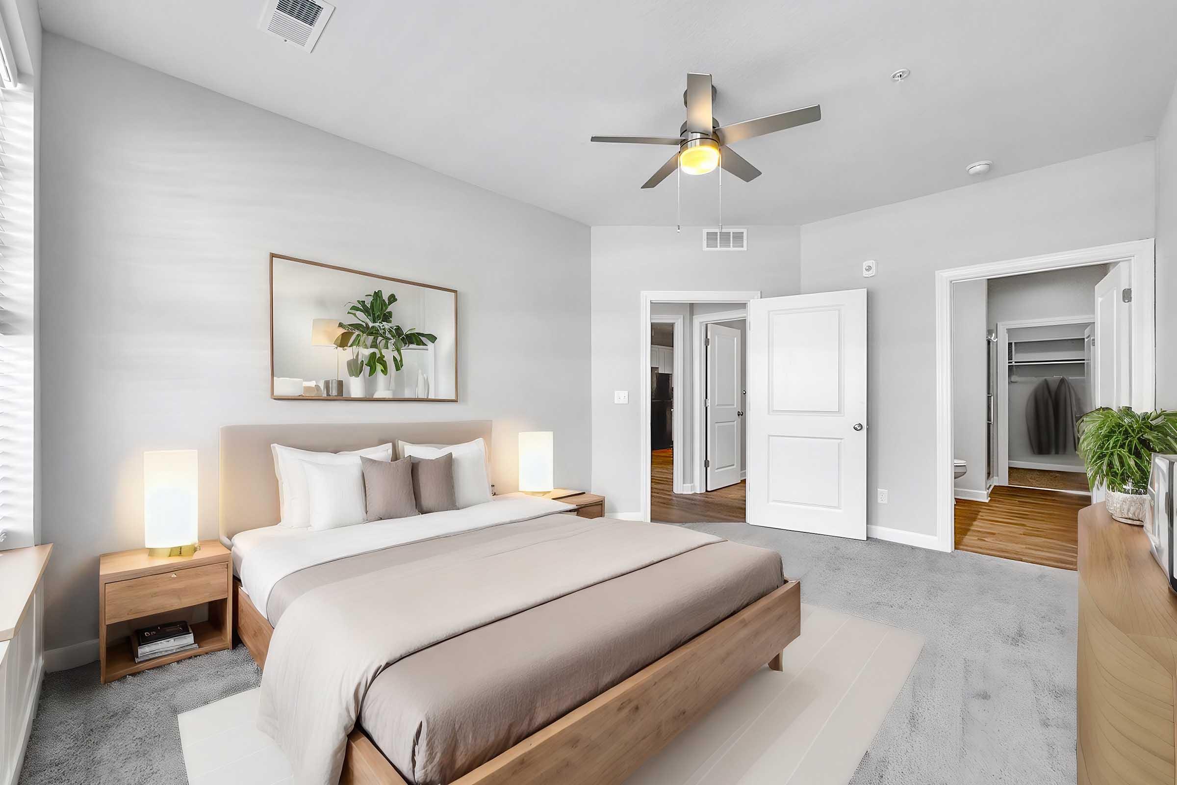 A modern bedroom featuring a light gray color scheme, a queen-sized bed with neutral bedding, two bedside lamps, and a wall-mounted mirror above the bed. There are large windows letting in natural light, a ceiling fan, a wooden dresser, and an open closet visible through a doorway.