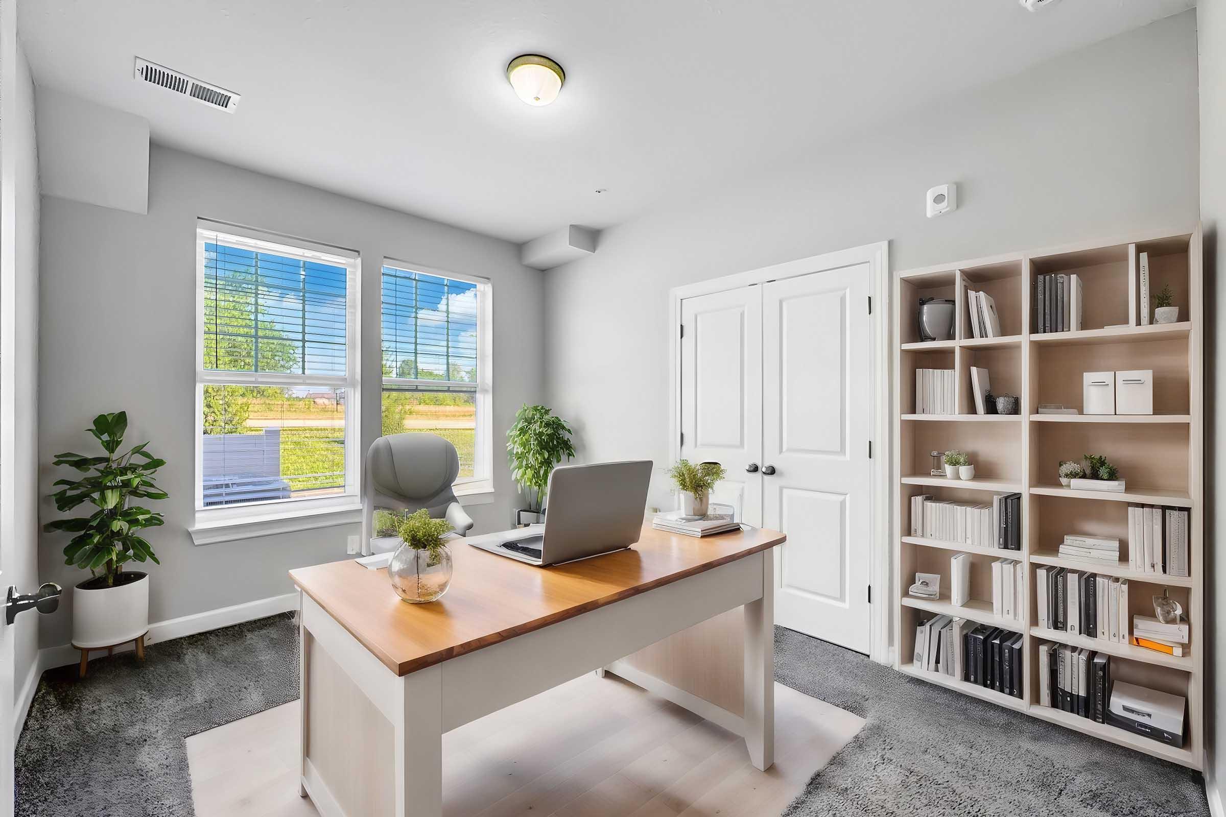 A modern home office featuring a wooden desk with a laptop, a comfortable chair, and a bookshelf filled with books and decorative items. Large windows allow natural light to enter, illuminating the room's gray walls and adding a touch of greenery with potted plants.
