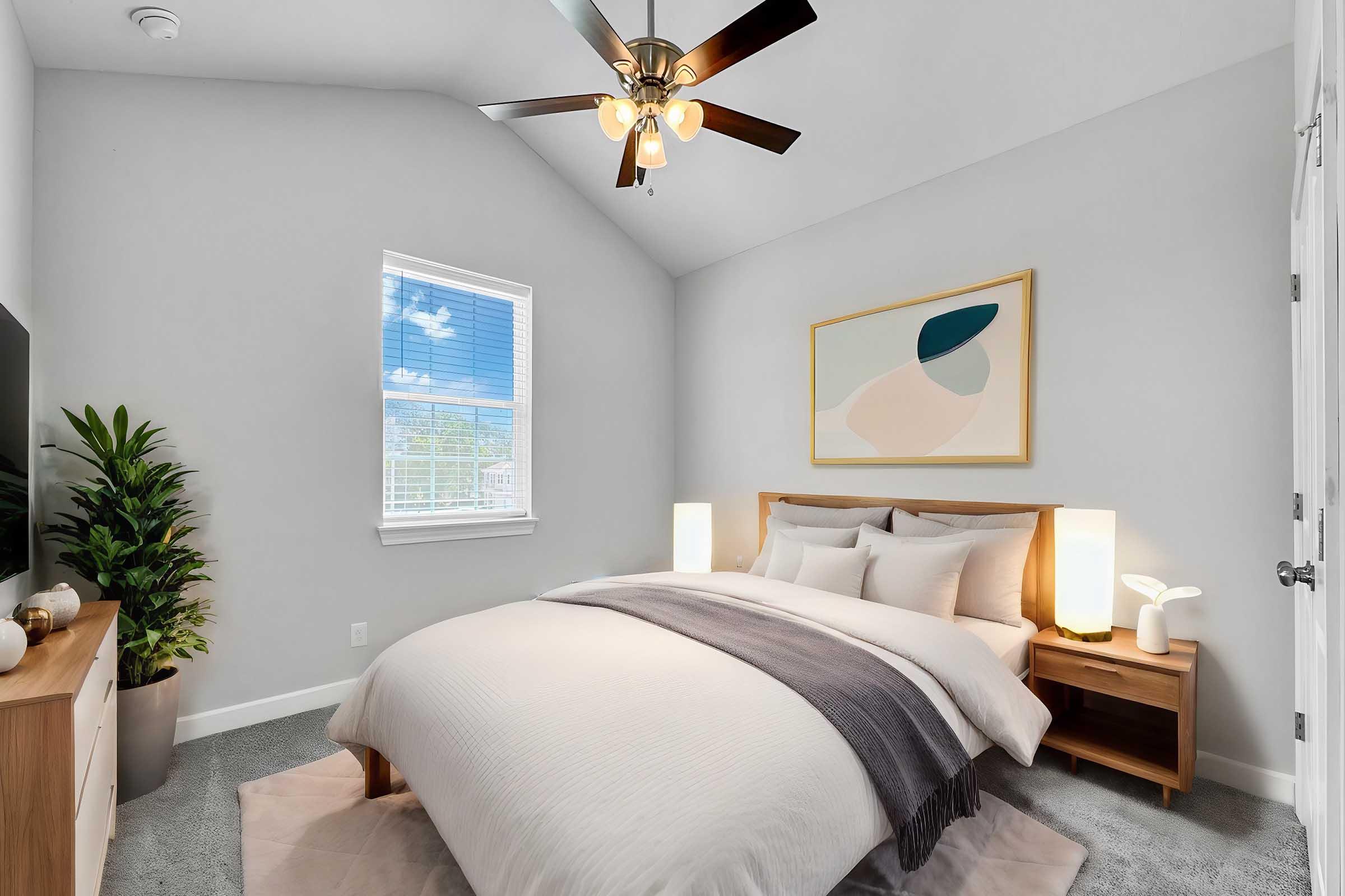 A cozy bedroom featuring a light gray color scheme, a comfortable bed with soft bedding, and a decorative throw. There are two bedside lamps, a wooden nightstand, and a modern painting on the wall. The large window lets in natural light, and a potted plant adds a touch of greenery to the space.