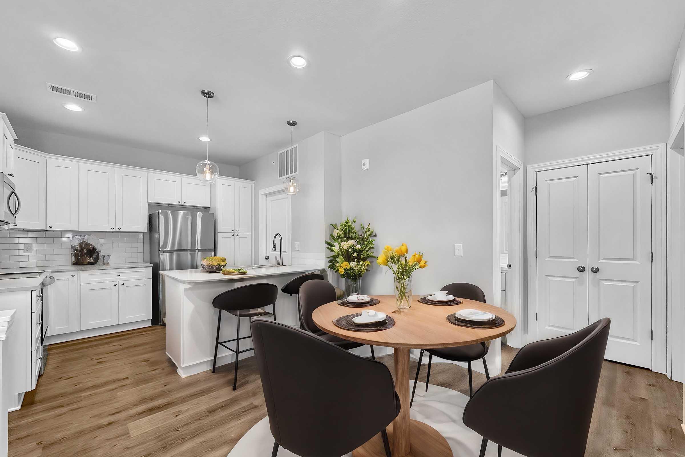 A modern kitchen with white cabinetry and stainless steel appliances. A wooden dining table with black chairs is set for four, adorned with floral arrangements. The space features pendant lighting, a large window, and an open layout that connects the kitchen to the dining area.