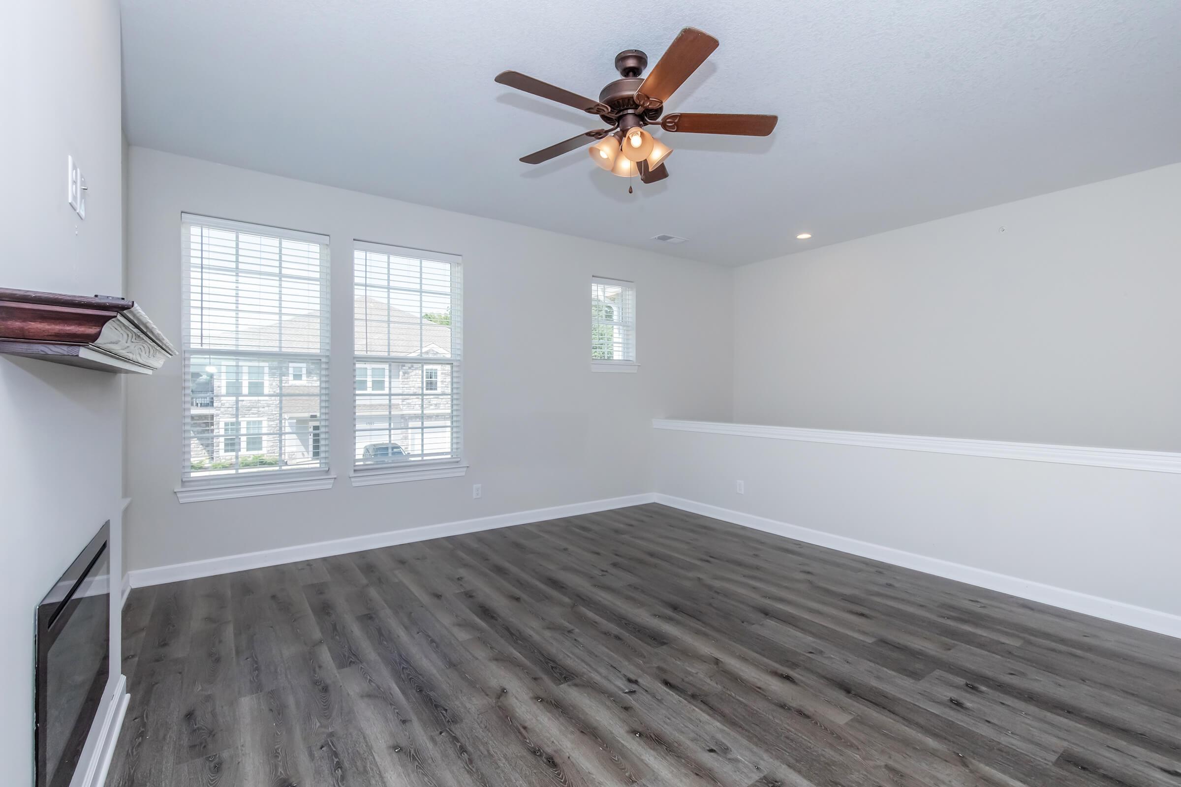 A spacious, modern living room featuring light-colored walls, a wooden floor, and a ceiling fan. It includes large windows with blinds allowing natural light and a small fireplace. The room is empty, creating a clean and inviting atmosphere.