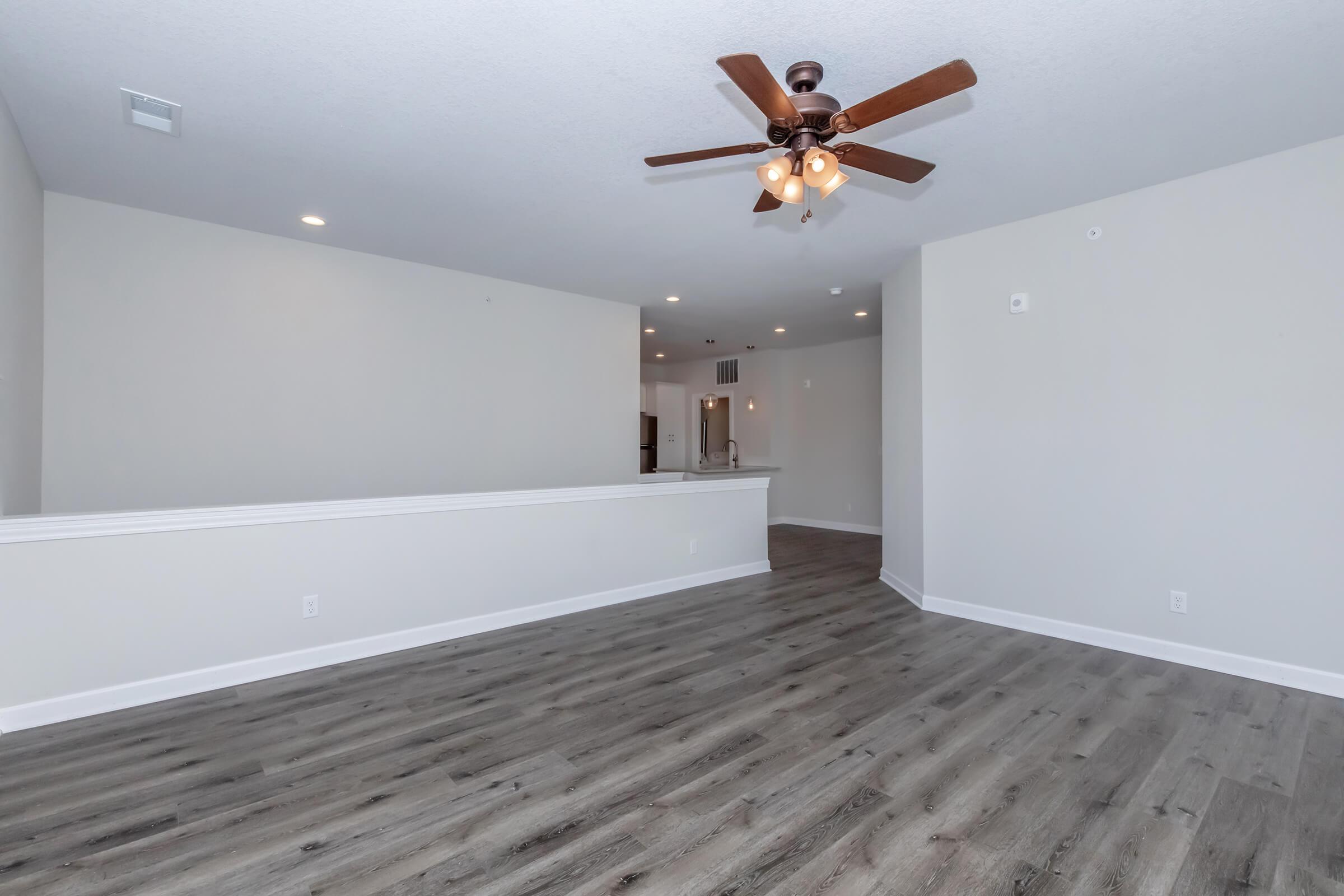 Spacious interior of a modern living room featuring light-colored walls and hardwood-style flooring. A ceiling fan with warm light fixtures hangs above, and there is an open space that leads to a kitchen area in the background. The ambiance is bright and inviting, suitable for various decorating styles.