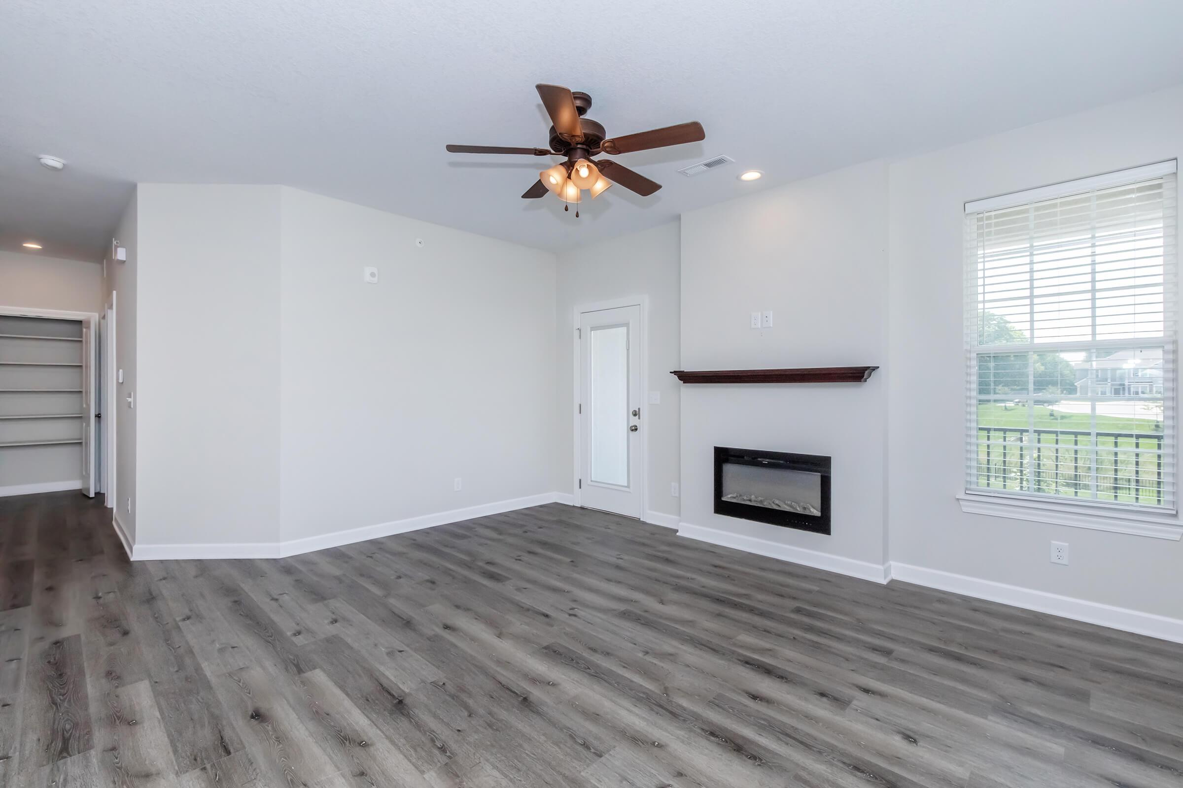A spacious, empty living room with light-colored walls and wooden flooring. It features a ceiling fan, a small electric fireplace in the corner, and large windows that provide natural light. There is a door leading outside, and a built-in shelf on the wall. The area is bright and inviting.