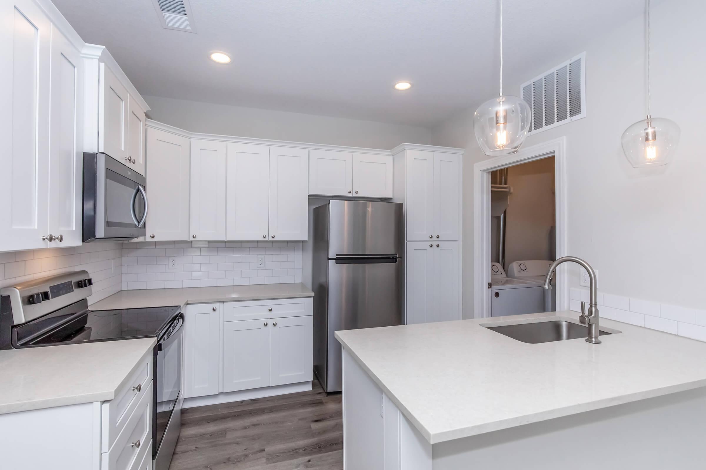 Modern kitchen featuring white cabinetry, stainless steel appliances, and a large countertop. The space includes a sink, a silver refrigerator, and a contemporary light fixture hanging above the island. Bright, neutral colors and laminate flooring create a clean, inviting atmosphere.