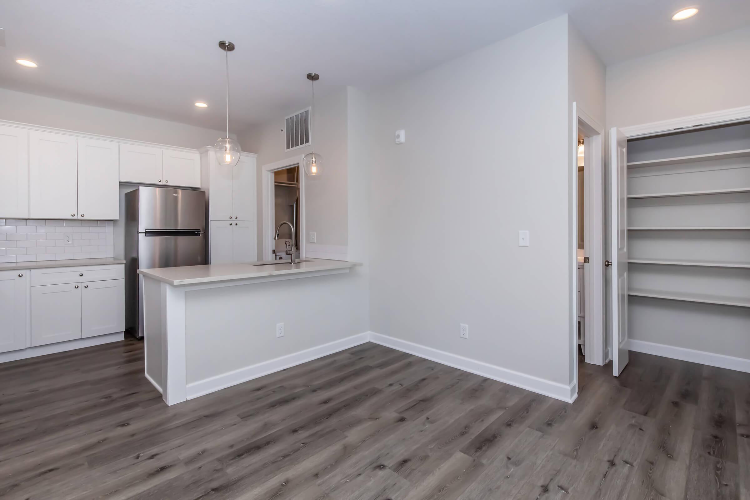 Modern kitchen and living space featuring white cabinetry, stainless steel appliances, and light wood flooring. The layout includes an island with a breakfast bar, pendant lighting, and an adjacent pantry. The walls are painted in a light color, creating an airy and open ambiance.