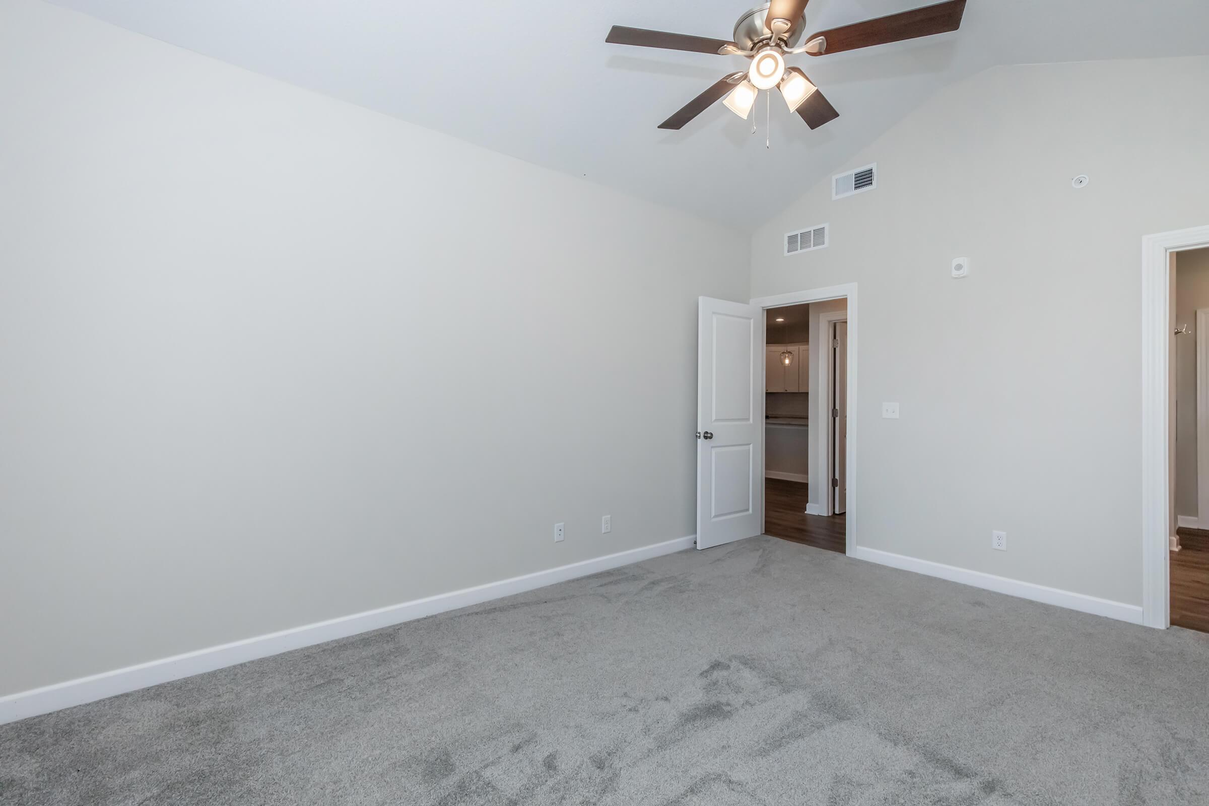 Empty room with light gray carpet, painted beige walls, and a ceiling fan. It features a doorway leading to another room and has no furniture. The ceiling is high with a sloped design, creating a spacious feel. Natural light comes from a window outside the frame.