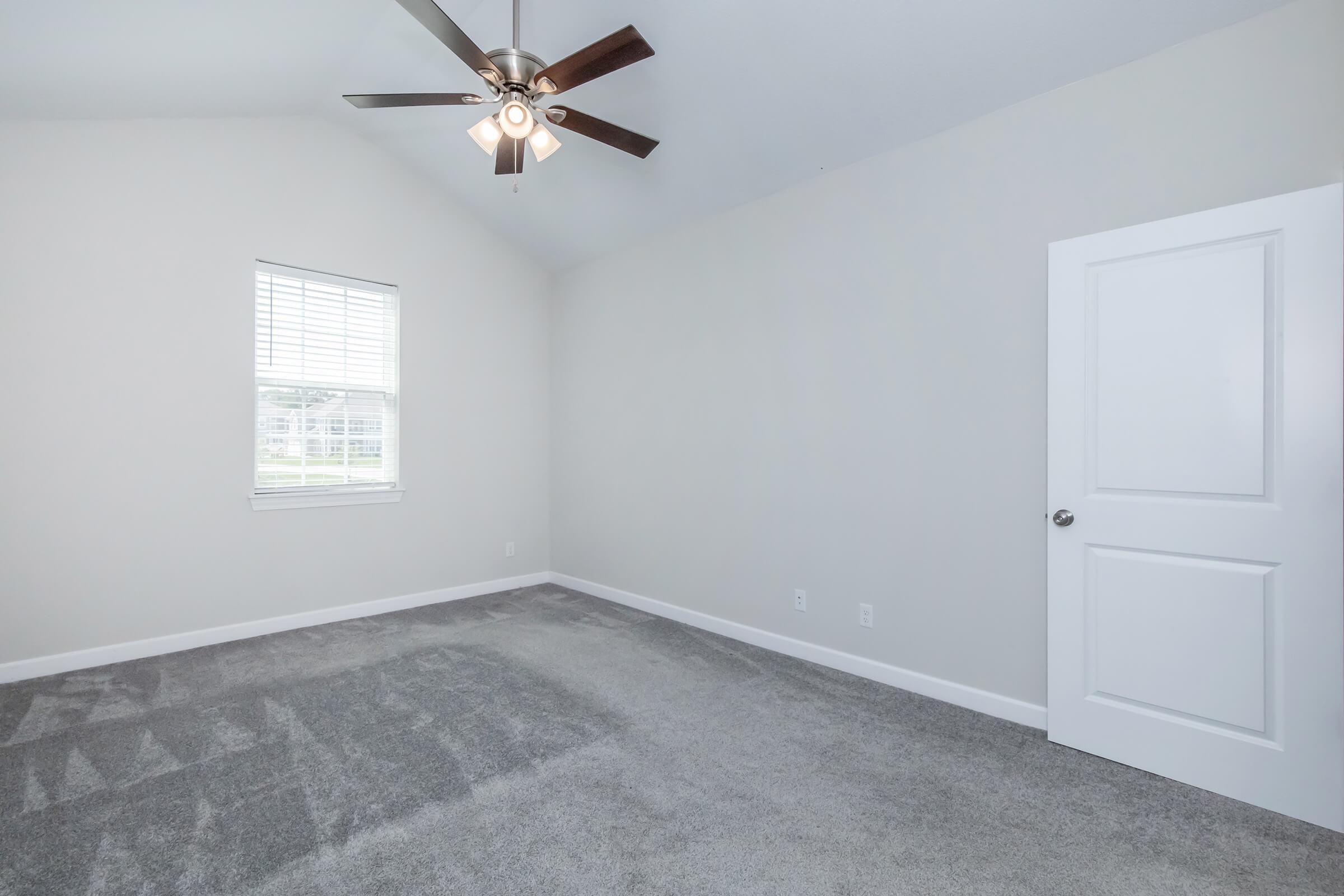 A spacious, empty room with a ceiling fan, high vaulted ceiling, and light gray carpet. It features a window letting in natural light, and a white door on the right wall. The walls are a soft neutral color, creating a bright and airy atmosphere ideal for decorating or furnishing.