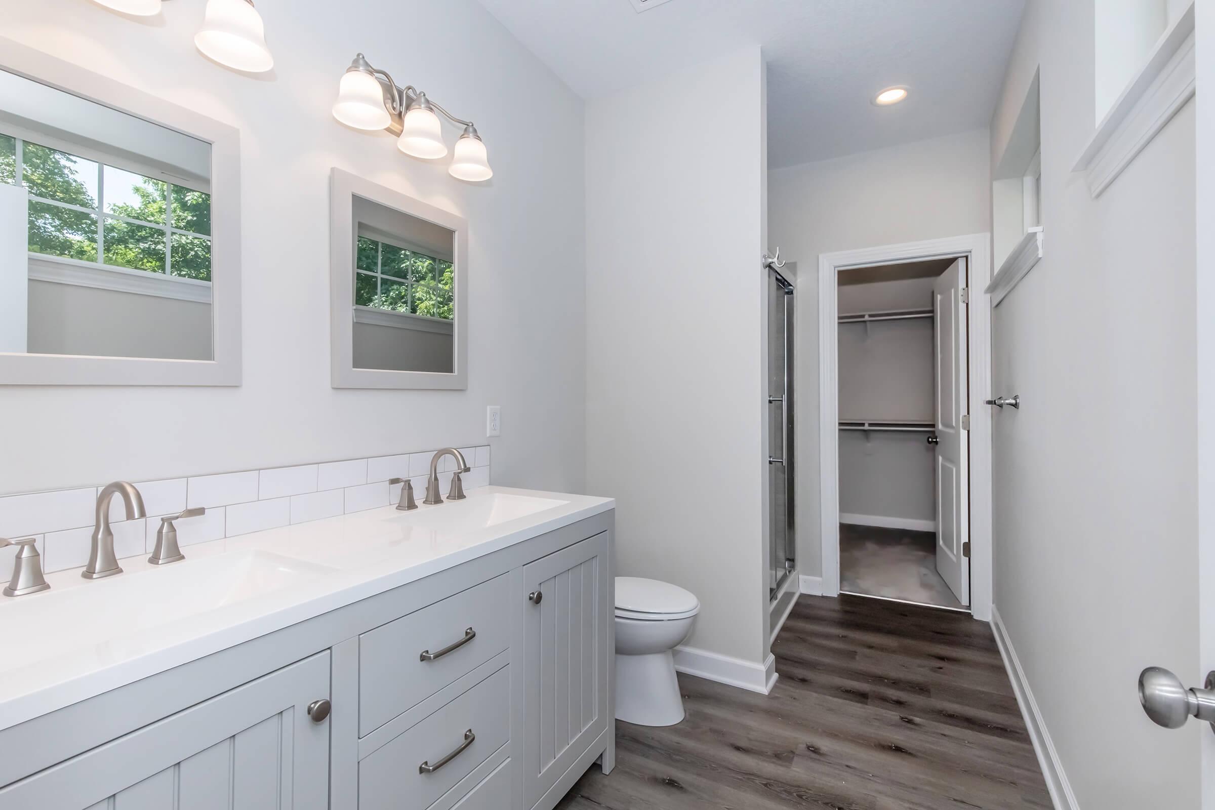 A modern bathroom featuring a double vanity with two mirrors, sleek light fixtures, and a neutral color palette. There's a white toilet and a door leading to a closet area. Natural light streams in from windows, illuminating the wood-look flooring.