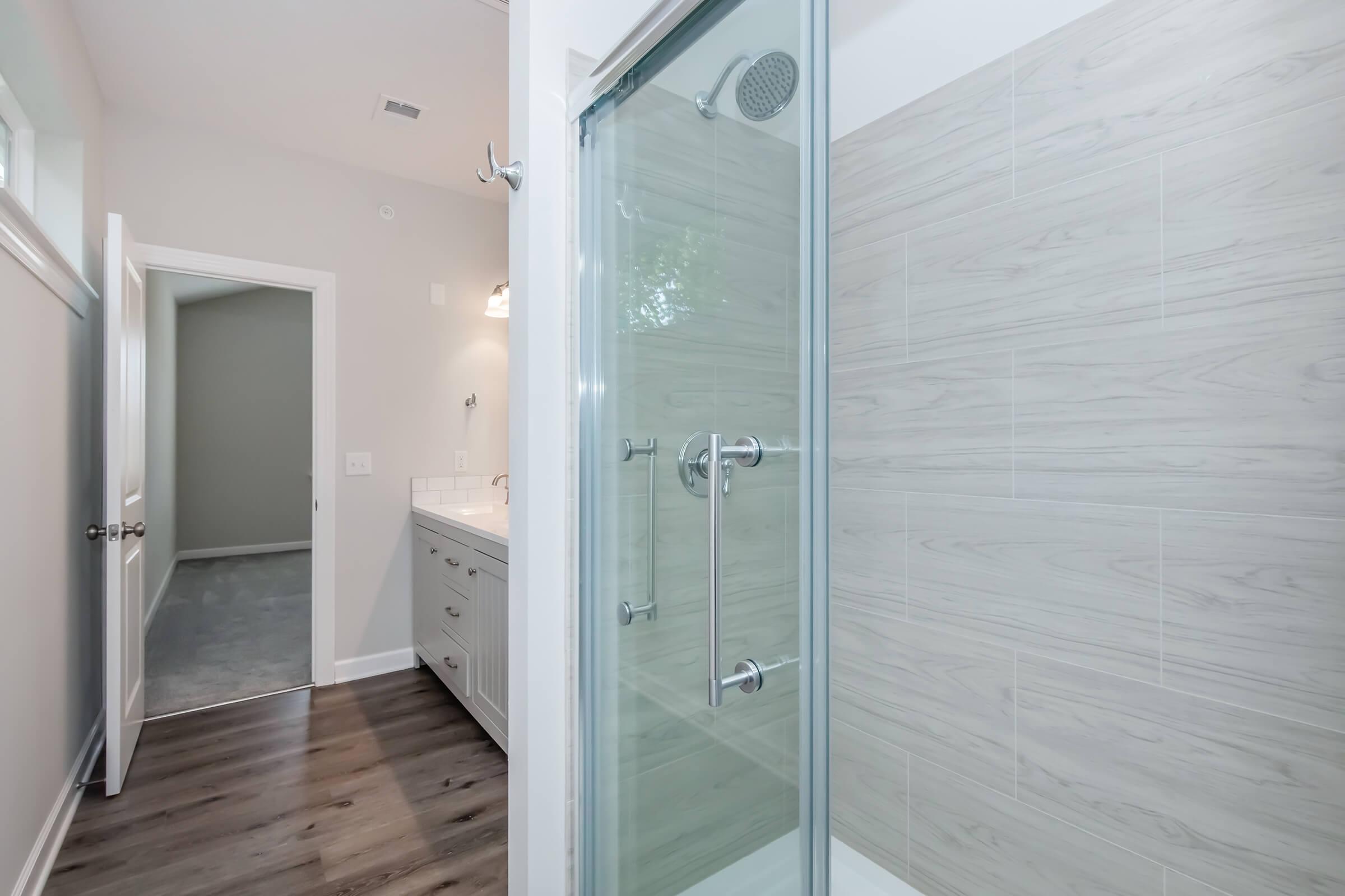 Modern bathroom interior featuring a glass-enclosed shower with chrome fixtures and a light wood tile backdrop. The adjacent room is partially visible with a neutral wall and door. The space is bright and airy with natural light coming through a window. The flooring is dark wood.