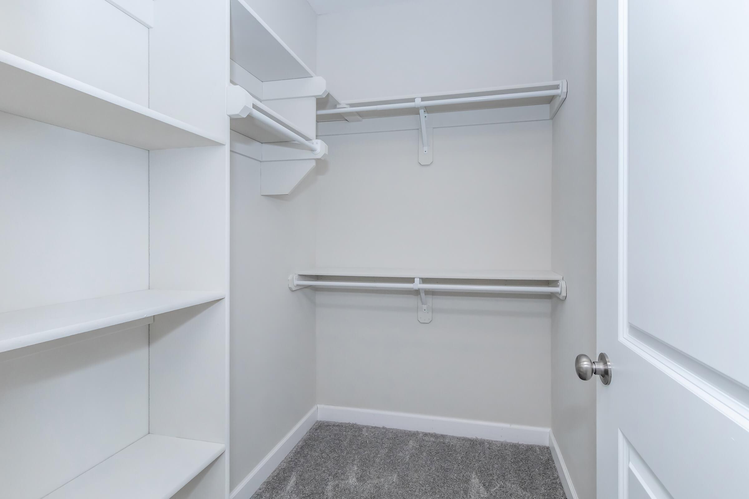 A spacious, empty closet featuring light gray walls, white shelving units on the left, and two hanging rods on the right. The floor is covered in light gray carpet, and a closed white door is visible on the right side. The overall look is clean and minimalistic.