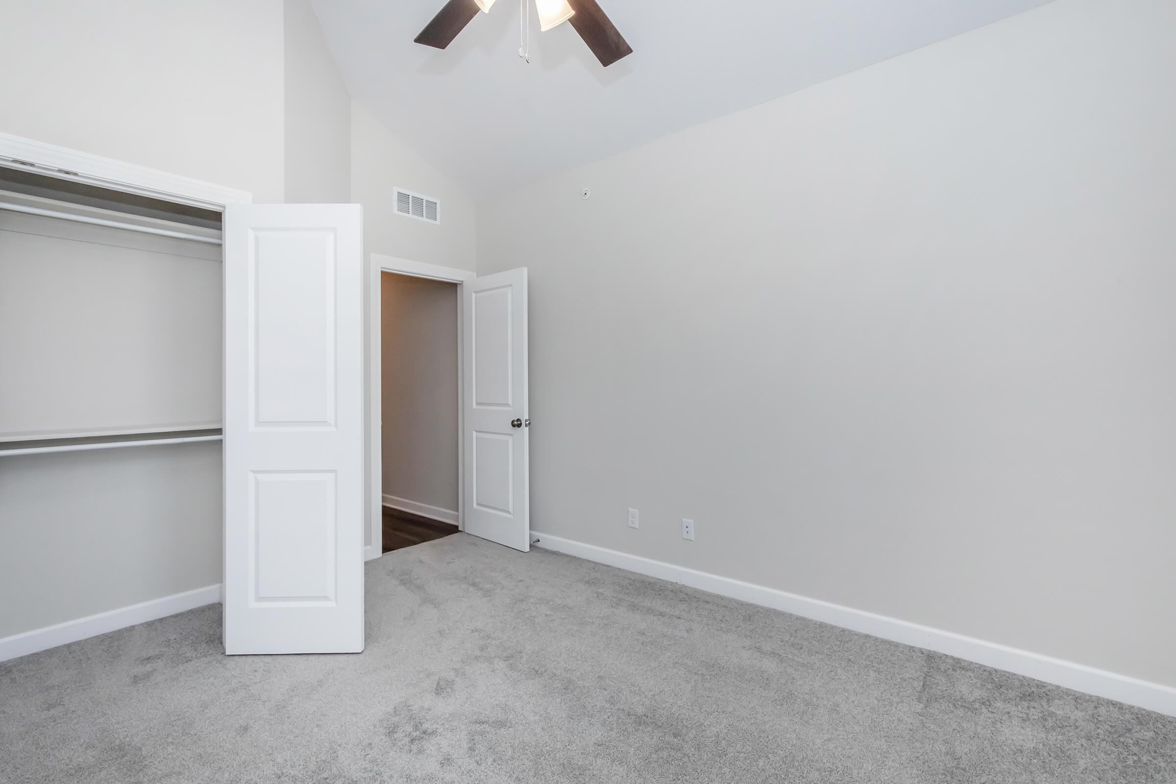 A spacious room featuring light gray carpet, a ceiling fan, and white walls. There are open double doors leading to a closet on the left, and another door at the far end that opens to a hallway. The room is well-lit with natural light.