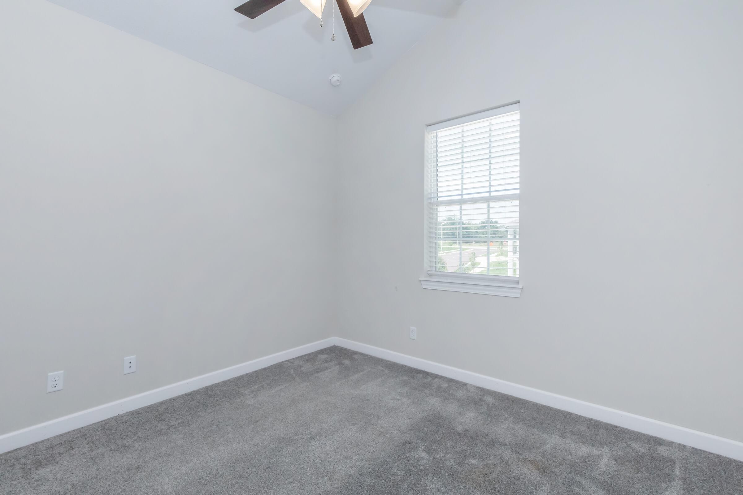 Empty room with light gray carpet and white walls, featuring a ceiling fan. A window with white blinds allows natural light to enter, revealing a view outside. The room has a vaulted ceiling and no furniture or decorations.