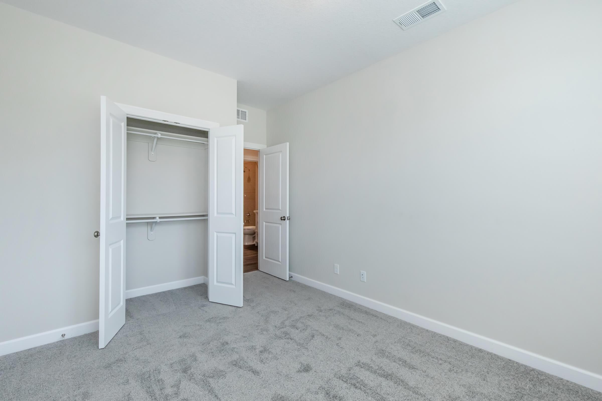 A vacant room with light gray carpet and beige walls. Two white closet doors are open, revealing an empty closet space. The room has a simple design with a light source coming from the doorway, leading to a bathroom area.
