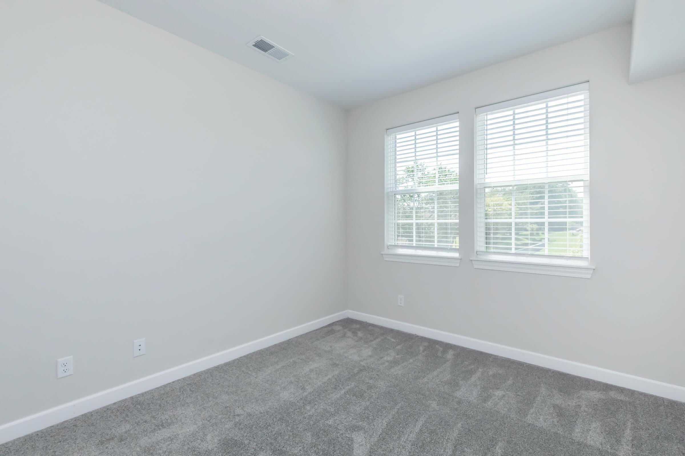 An empty room with light gray carpet and pale walls. The room features two windows with white blinds, allowing natural light to enter. There are minimalistic electrical outlets and no furniture, creating a spacious and clean aesthetic.