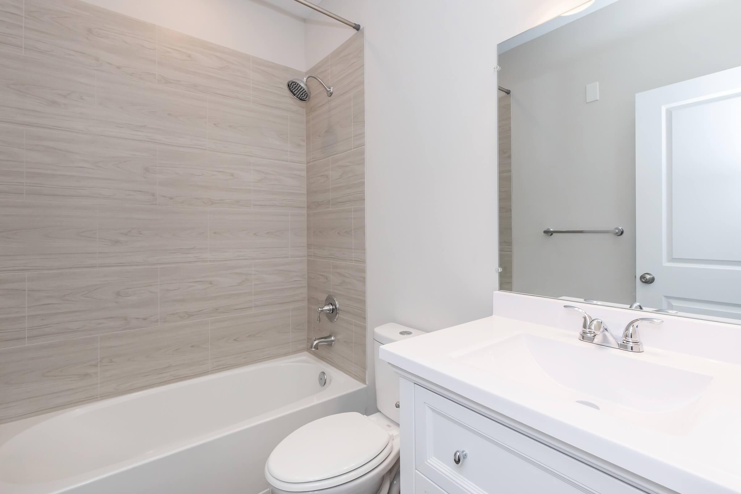 A modern bathroom featuring a white bathtub with a showerhead, light-colored tiled walls, a white vanity with two sinks, and a large mirror. The space is well-lit and has a clean, minimalist design. A towel rod is mounted on the wall, and a door is visible in the background.