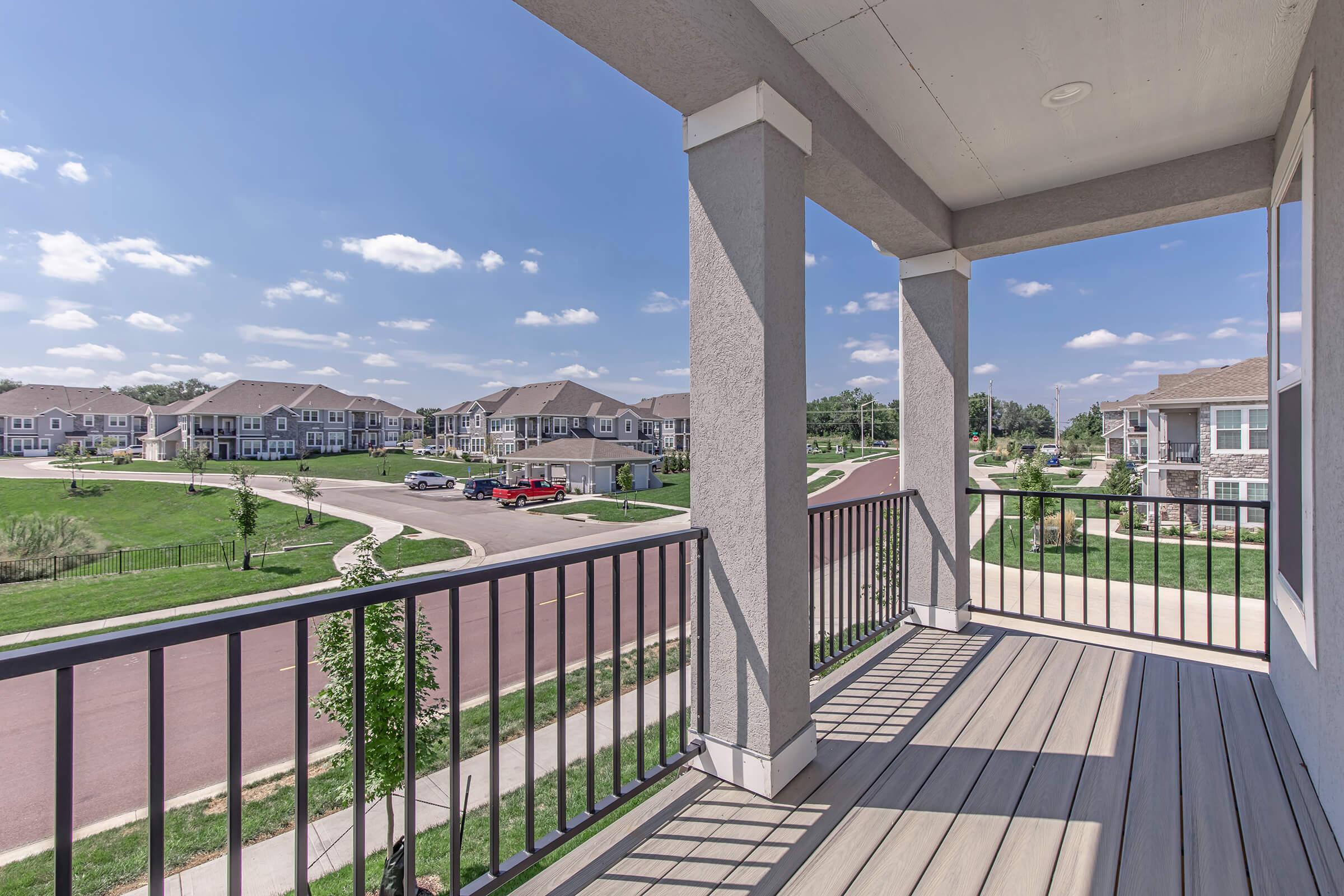 View from a balcony showcasing a sunny neighborhood with multiple residential buildings, a curved road, and parked vehicles. The scene features clear blue skies and scattered clouds, highlighting the well-maintained surroundings and inviting atmosphere.