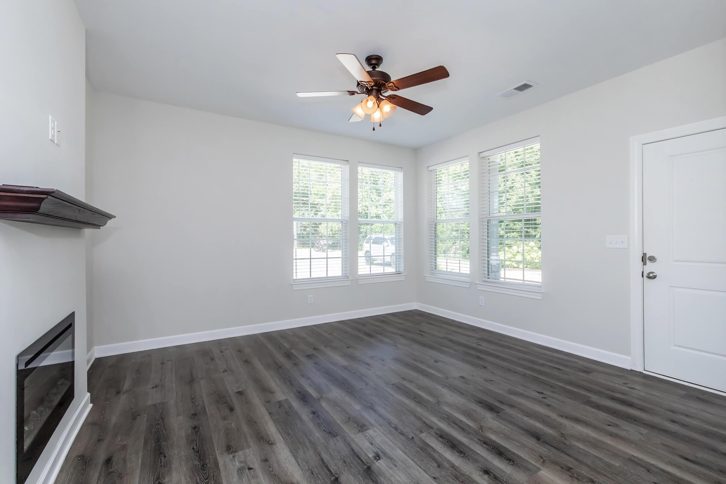 A bright, open living room featuring light-colored walls, large windows with white blinds, and a ceiling fan. The floor is covered with dark wood-like flooring, and there is a modern, unlit fireplace on one wall. The entrance door is visible on the right side.