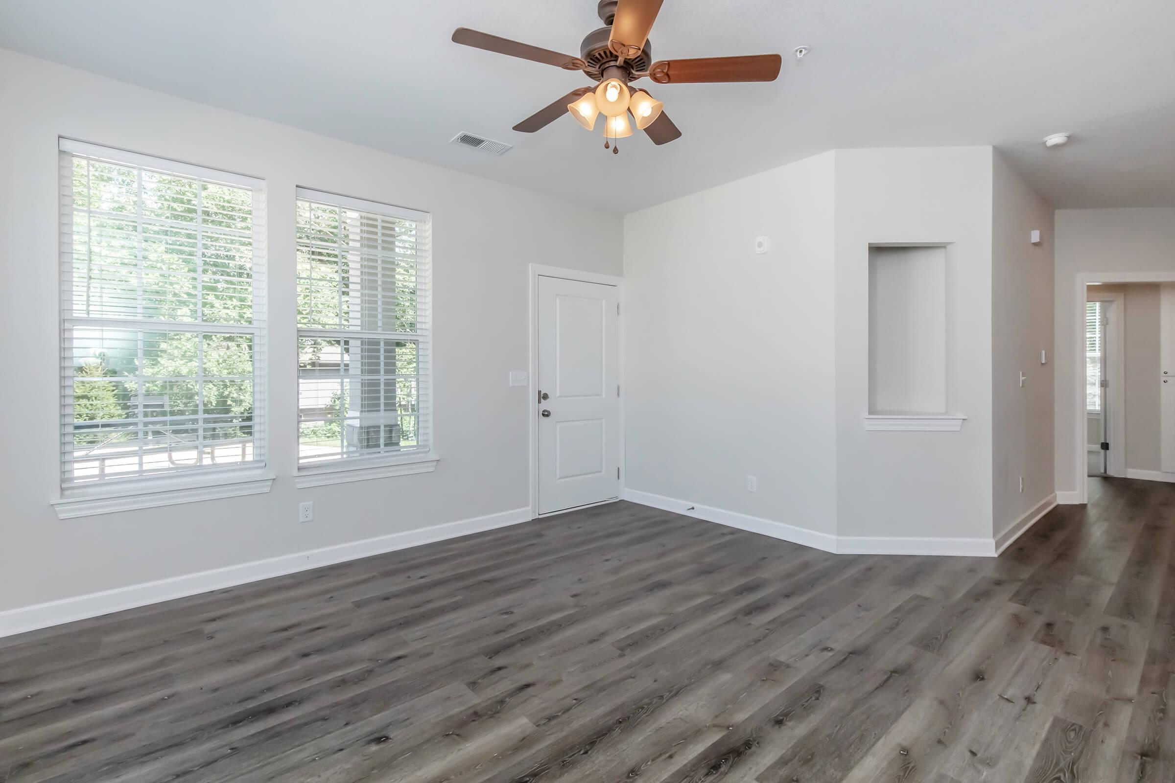 A bright, airy living room featuring large windows that allow natural light to flood in. The room has light-colored walls and hardwood flooring, complemented by a ceiling fan with wooden blades. A door leads to the outside, while a doorway leads to another room in the background.