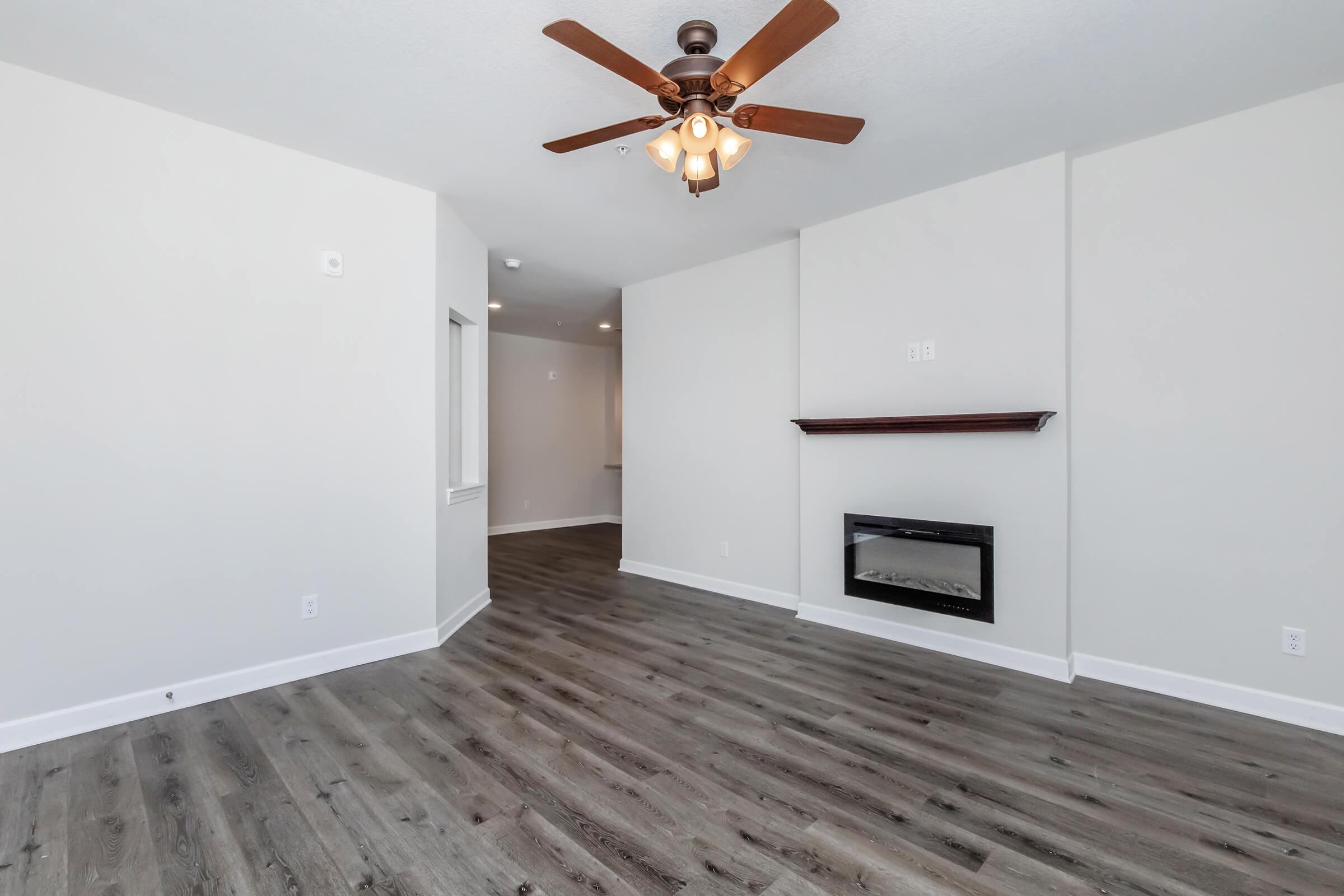 A spacious, empty living room featuring light-colored walls, warm wood laminate flooring, a ceiling fan with light fixtures, and a modern electric fireplace. An adjoining area is visible in the background, suggesting an open concept layout.