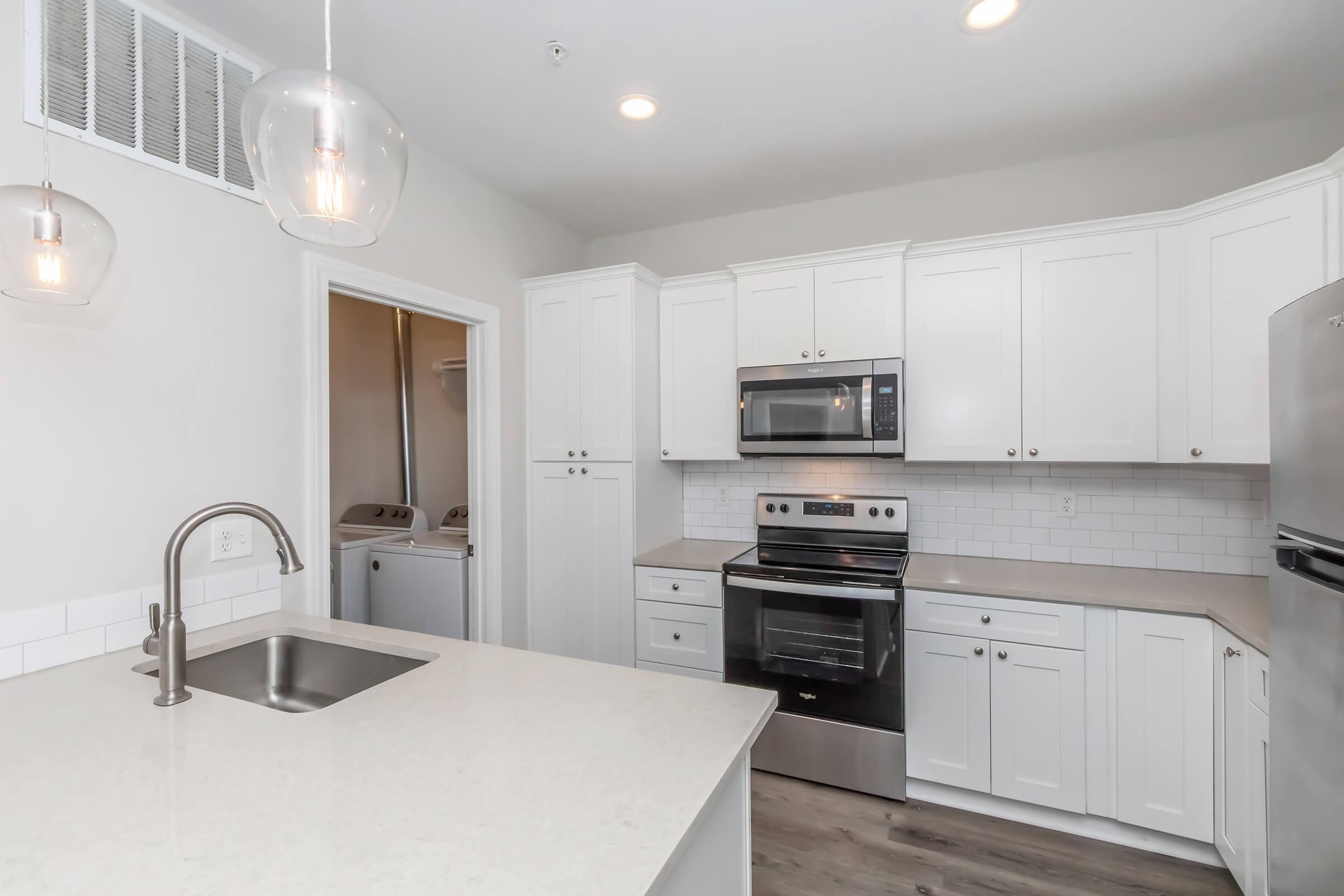 A modern kitchen featuring white cabinetry, stainless steel appliances including an oven and microwave, a light-colored countertop, and pendant lighting. There is a sink in the foreground and a laundry area visible through an open door. The overall design is bright and contemporary.
