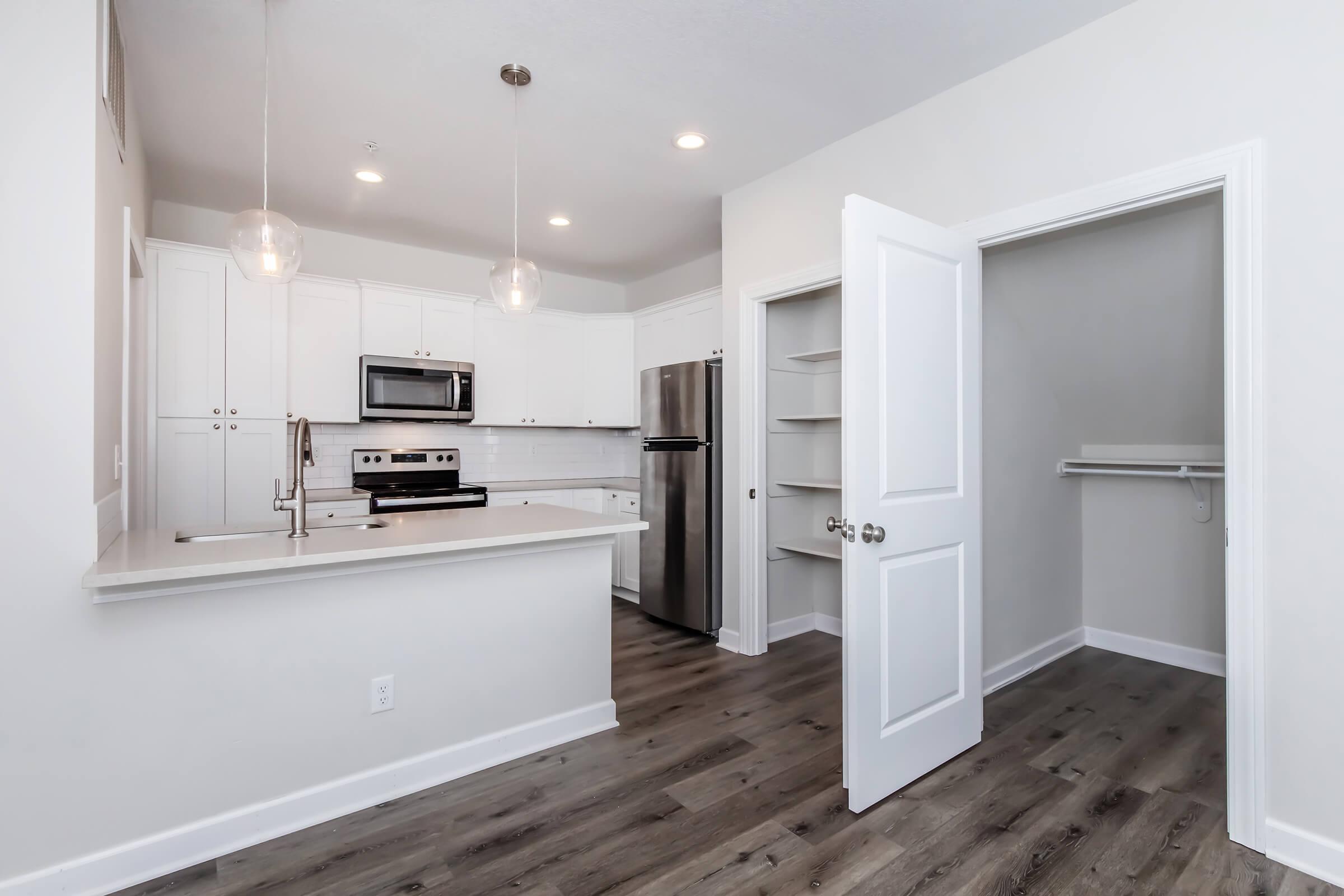 Modern kitchen with white cabinets, stainless steel appliances, and a kitchen island. Open layout leads to a pantry closet with shelves. The space features gray wood-like flooring and recessed lighting, creating a bright and inviting atmosphere.