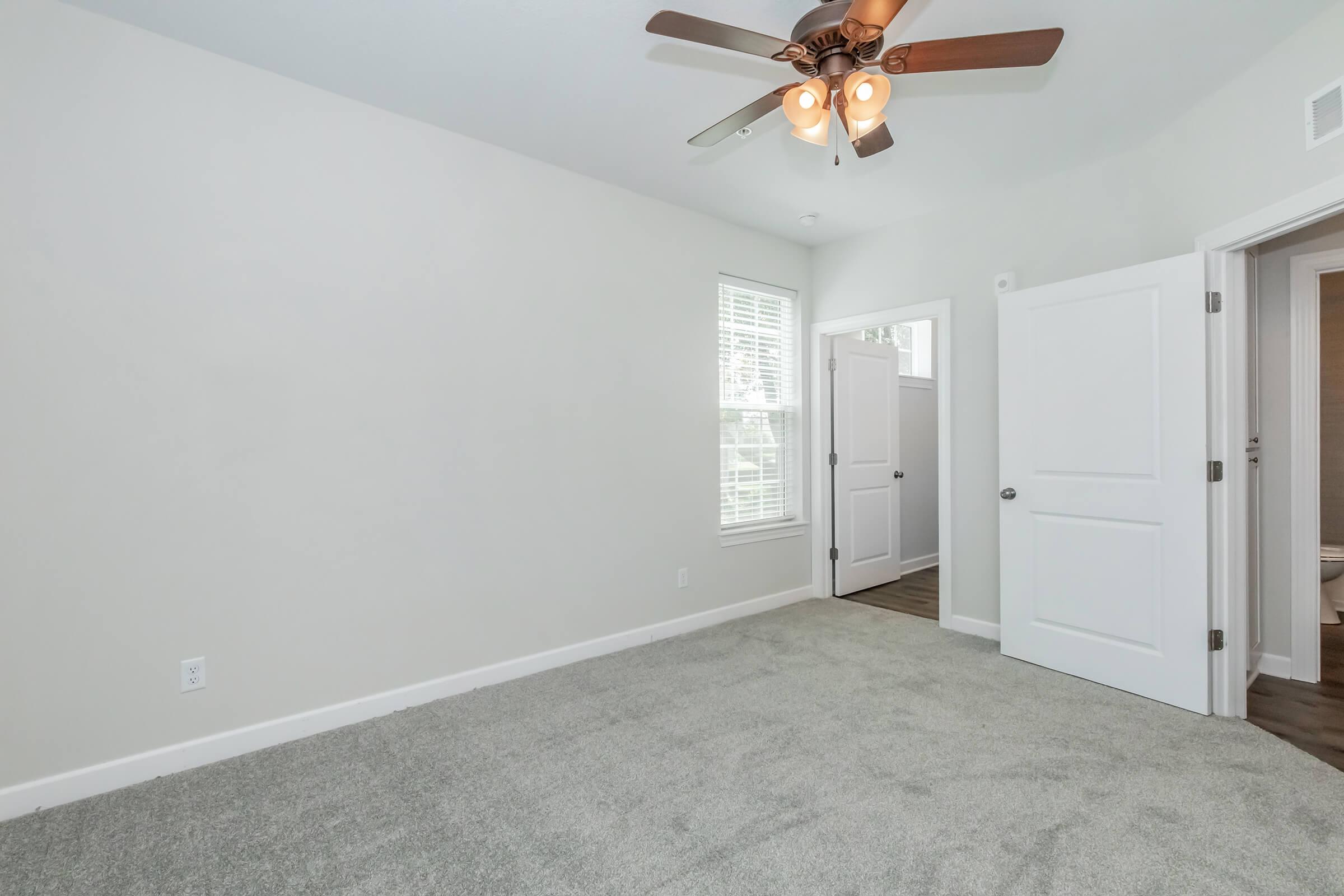 A bright, empty room with light gray carpet and white walls. A ceiling fan with light fixtures hangs above, and a window provides natural light. There is a partially open white door leading to another room, suggesting a modern and spacious layout.