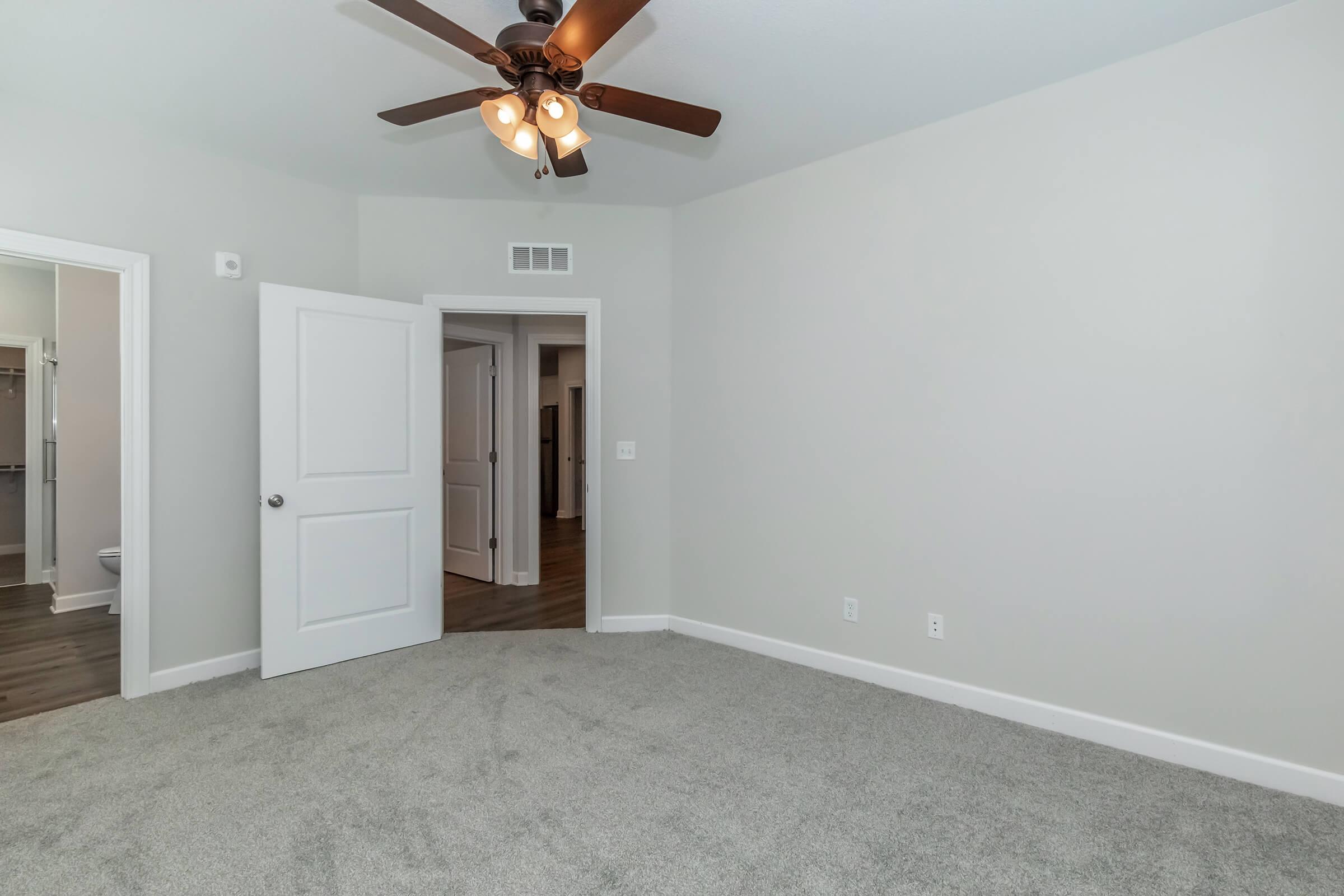 A spacious room with light gray walls and carpet flooring. It features a ceiling fan with four lights and a wide doorway leading to another area. A bathroom and an additional room are visible through open doors on the left. The overall ambiance is bright and inviting.