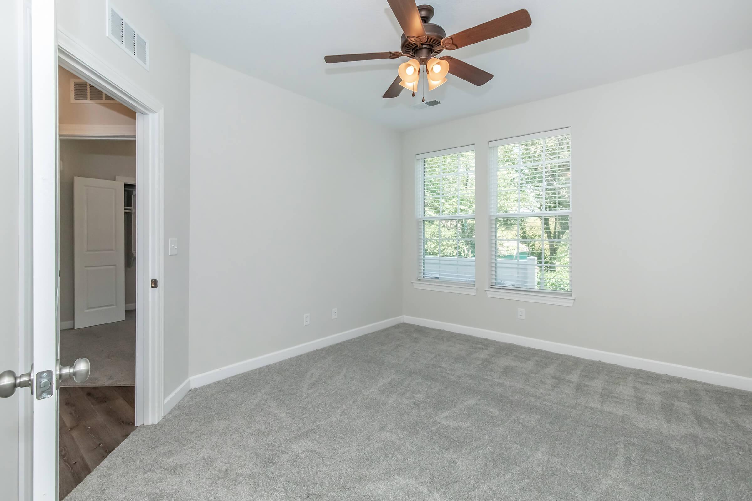 A well-lit, empty bedroom featuring light grey carpet, a ceiling fan with wooden blades, and two windows with white blinds. The walls are painted a soft neutral color. A doorway leads to another room, and the overall atmosphere is bright and spacious.