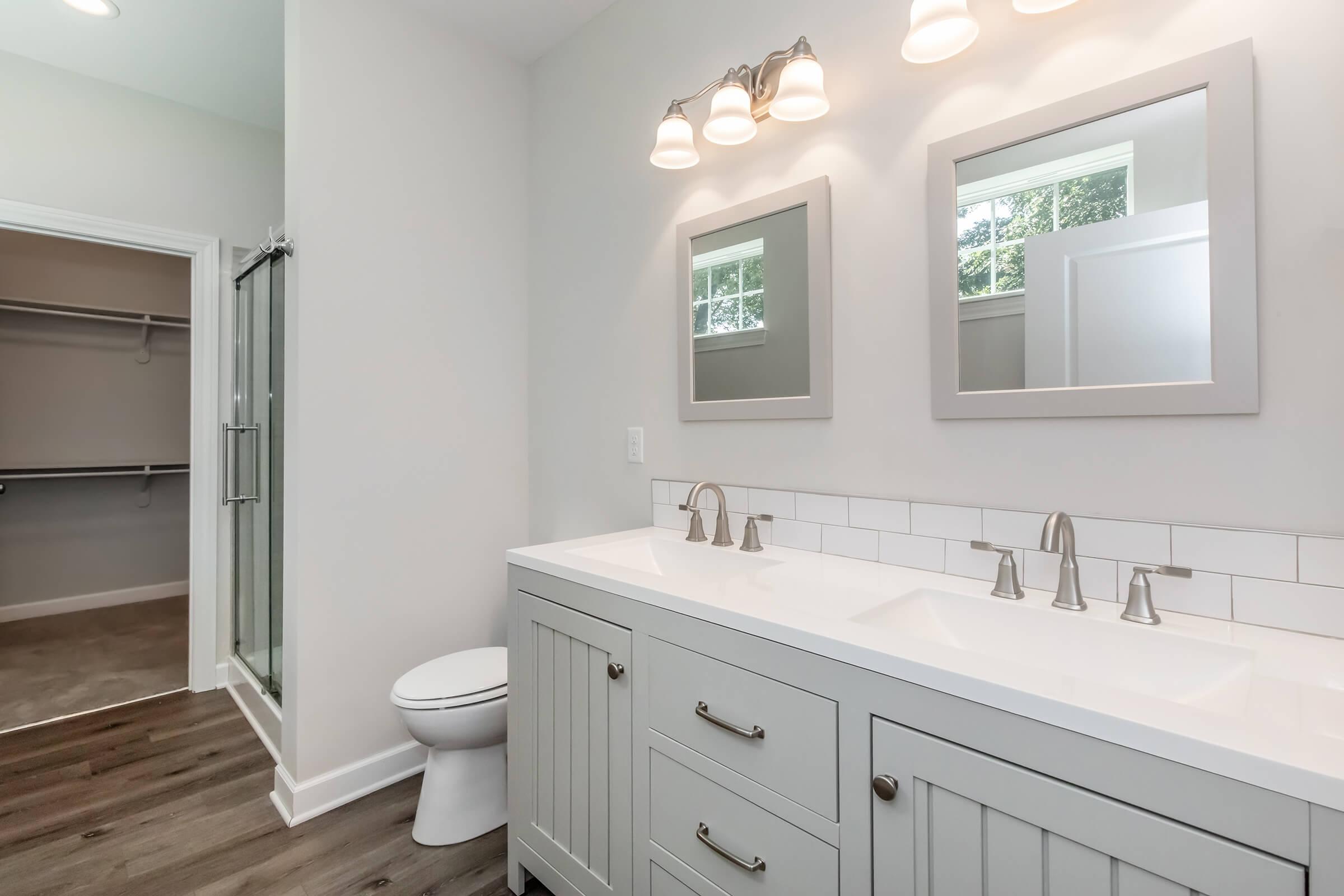 A modern bathroom featuring a double vanity with two sinks, two mirrors, and stylish light fixtures. The walls are painted a light color, and there is a glass shower stall on one side. A toilet is placed beside the vanity, and a spacious walk-in closet is visible in the background. The flooring is a natural wood finish.